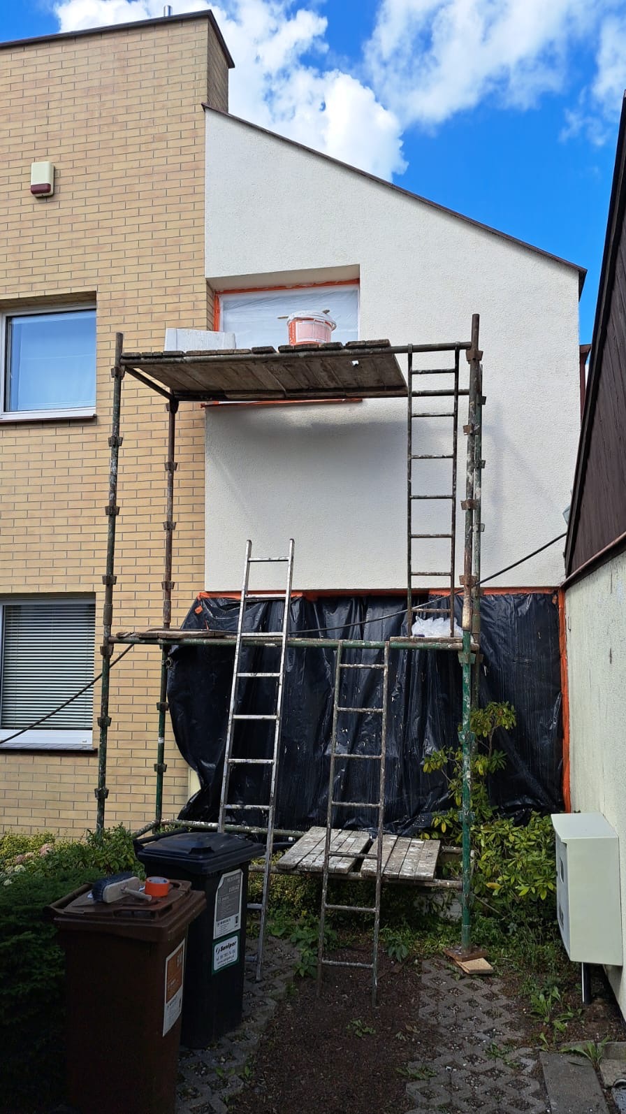 Prace elewacyjne w toku: rusztowanie przy budynku z częściowo otynkowaną i pomalowaną elewacją, widoczne zabezpieczenia okna i wiadro z farbą na rusztowaniu, obok śmietniki i drabiny.