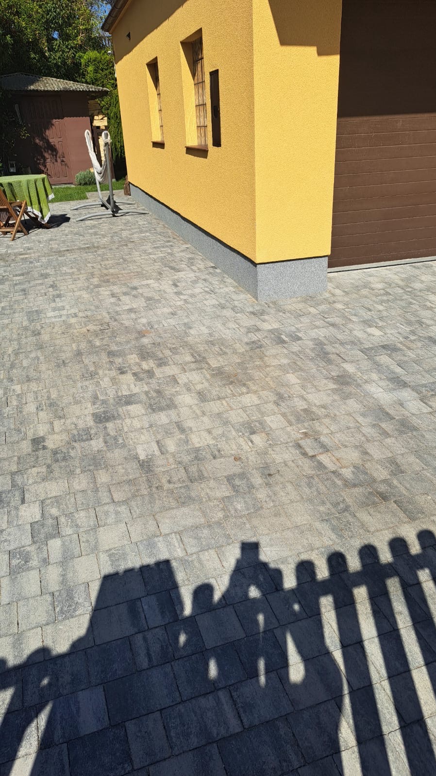 Ułożona kostka brukowa w odcieniach szarości przed żółtą elewacją budynku z brązową bramą garażową, cień osoby robiącej zdjęcie i cień ogrodzenia.