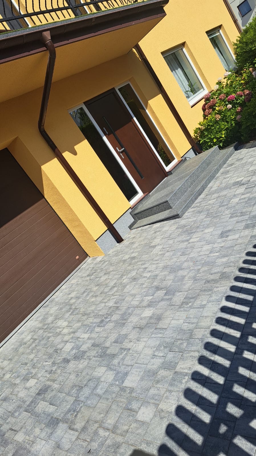 Żółty dom jednorodzinny z brązowymi drzwiami i bramą garażową, szara kostka brukowa na podjeździe, schody wejściowe z szarego kamienia, krzewy hortensji.