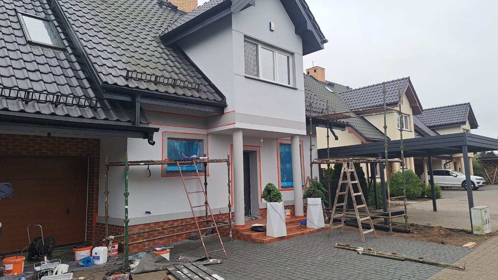 Dom w trakcie prac elewacyjnych: szara fasada z pomarańczowymi akcentami wokół okien i kolumn, okna zabezpieczone folią, rusztowania, kostka brukowa na podjeździe.