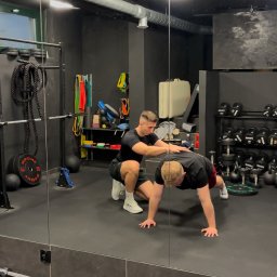 Dominik Kokoszko - Trener personalny pomaga mężczyźnie w ćwiczeniach pompek w siłowni z lustrami, widoczne ciężary, liny i gumy oporowe.