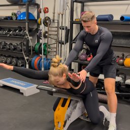 Dominik Kokoszko - Trener personalny asystuje kobiecie podczas ćwiczenia na ławce w siłowni, dbając o prawidłową postawę i technikę. W tle widoczne sprzęty do ćwiczeń.