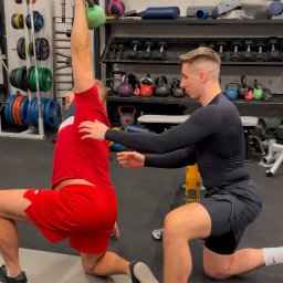 Dominik Kokoszko - Trener asekuruje ćwiczącego z zielonym kettlebell nad głową, w pozycji wykroku na macie w siłowni. Widoczny sprzęt do ćwiczeń na półkach w tle.