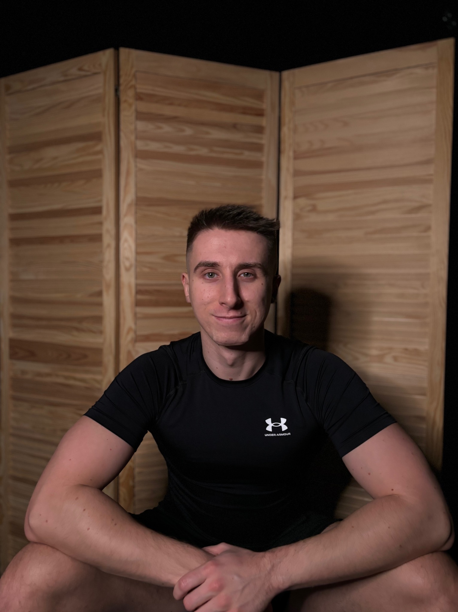 Młody mężczyzna w czarnej koszulce sportowej z logo Under Armour, siedzący na tle drewnianego parawanu.