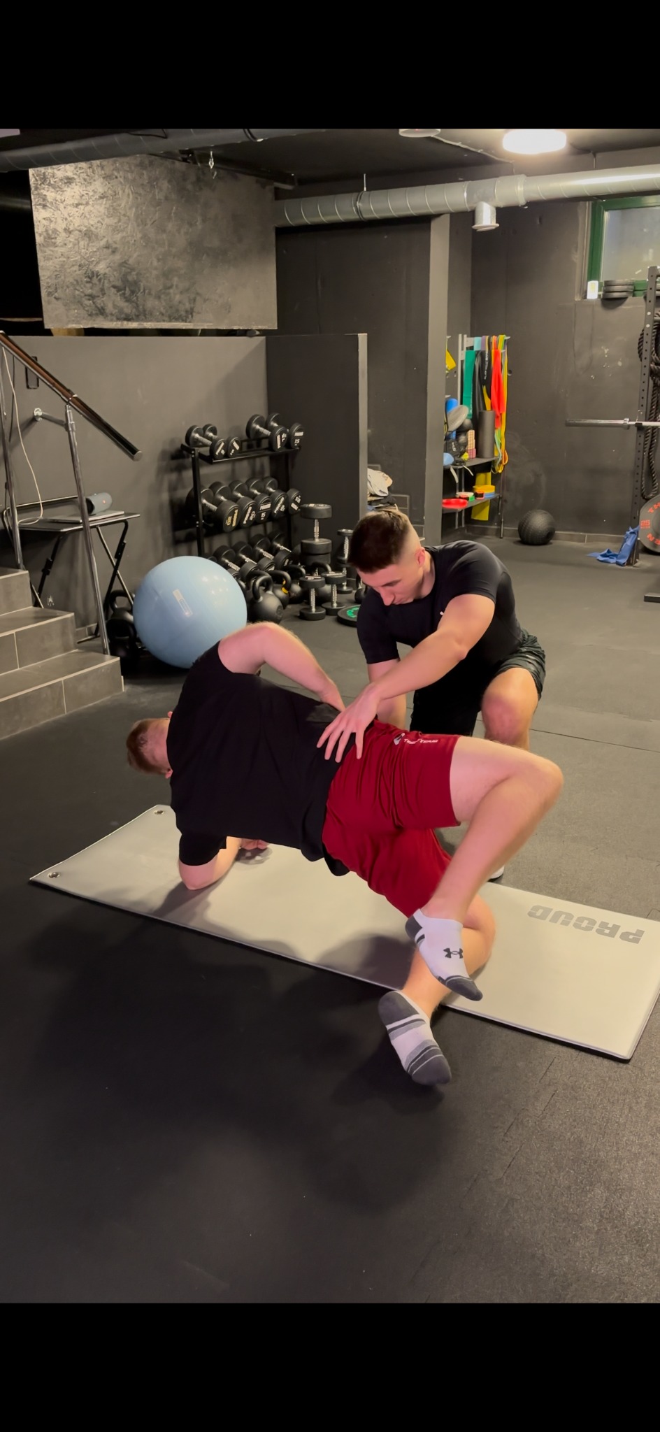 Trener personalny koryguje pozycję klienta podczas ćwiczenia na macie w siłowni z widocznymi hantlami i drabinkami.