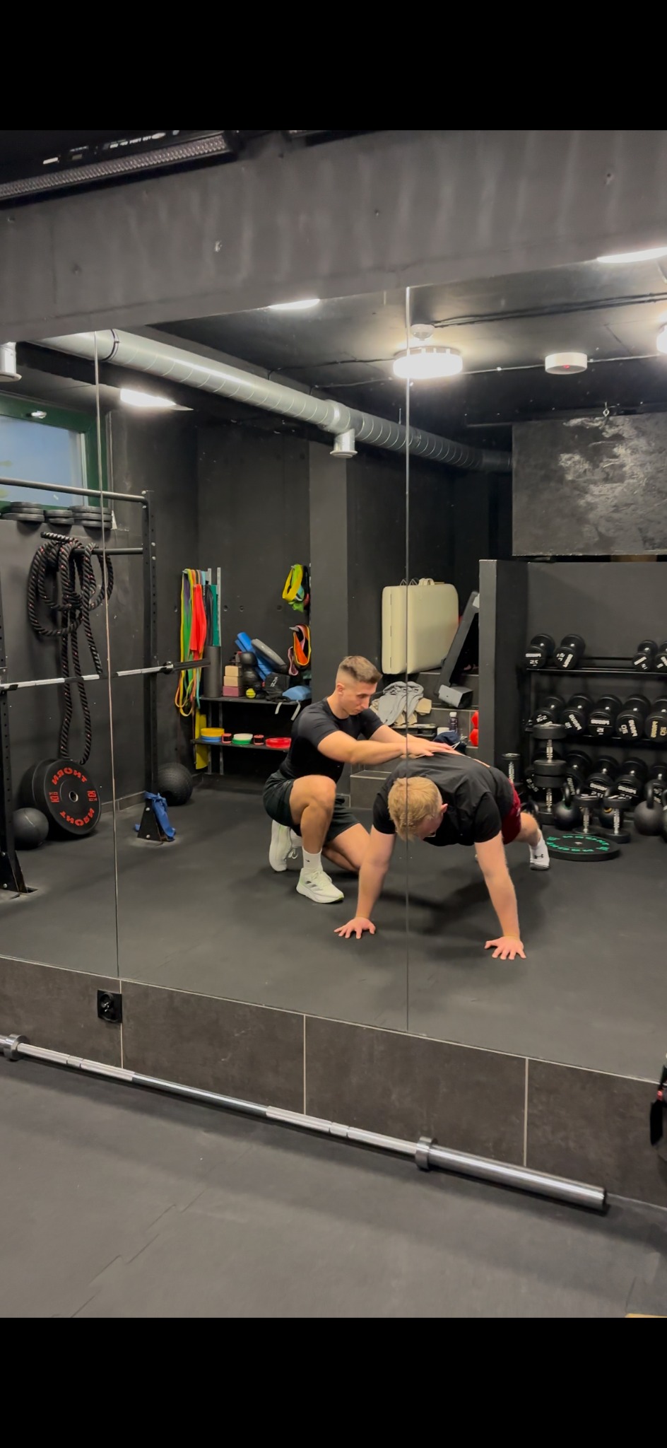 Trener personalny pomaga mężczyźnie w ćwiczeniach pompek w siłowni z lustrami, widoczne ciężary, liny i gumy oporowe.