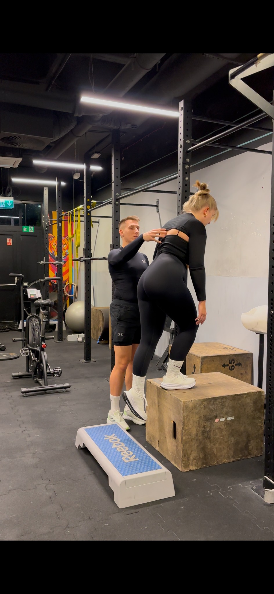 Trener personalny asekuruje kobietę w czarnym stroju podczas wchodzenia na skrzynię w siłowni. W tle widoczny sprzęt do ćwiczeń i kolorowe dekoracje.