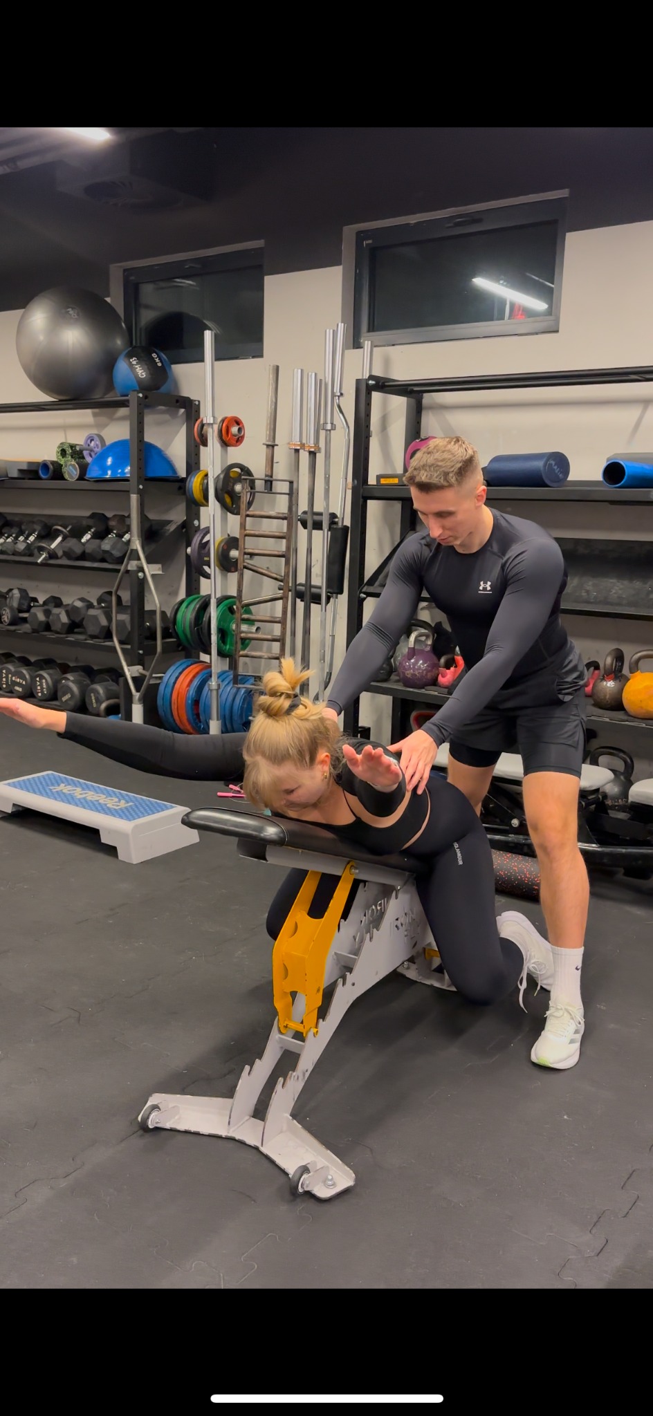 Trener personalny asystuje kobiecie podczas ćwiczenia na ławce w siłowni, dbając o prawidłową postawę i technikę. W tle widoczne sprzęty do ćwiczeń.