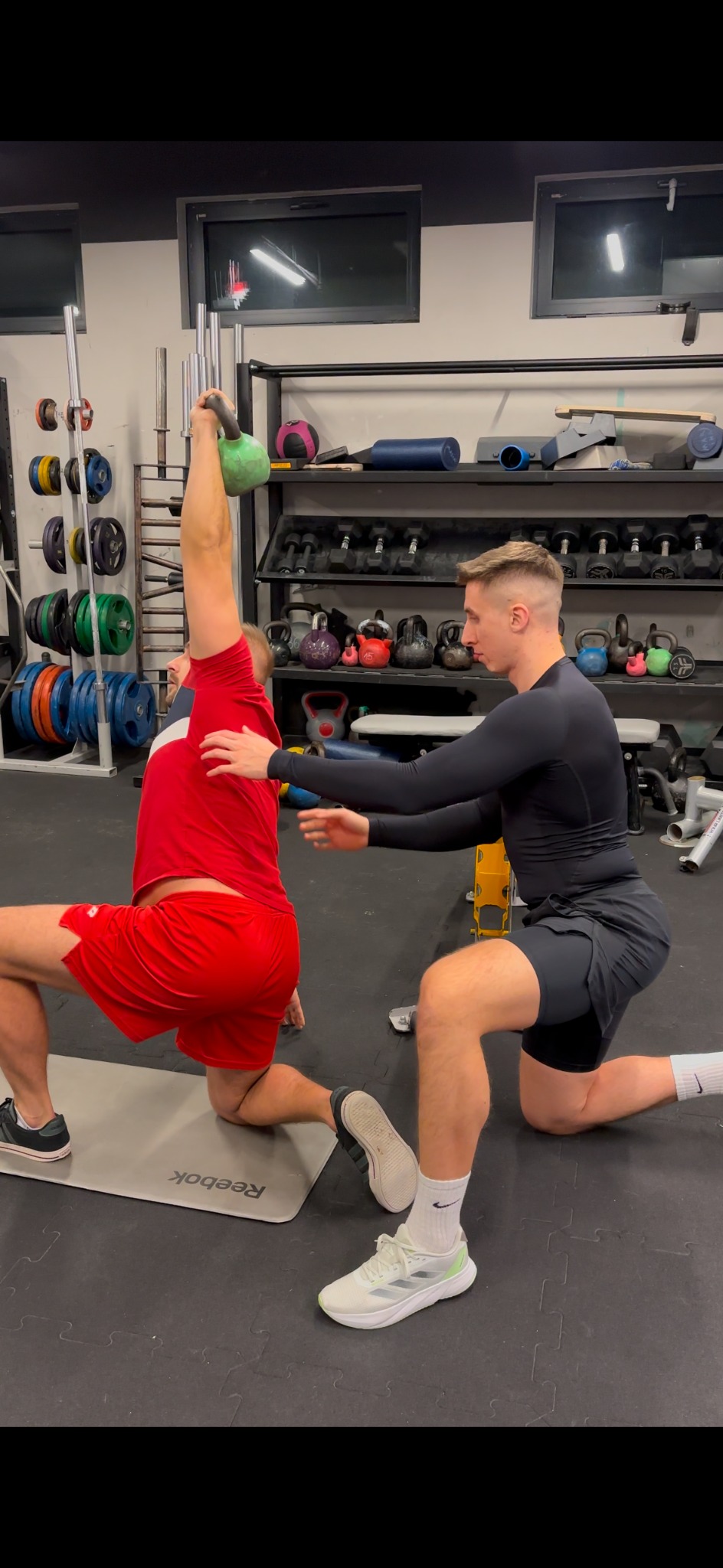 Trener asekuruje ćwiczącego z zielonym kettlebell nad głową, w pozycji wykroku na macie w siłowni. Widoczny sprzęt do ćwiczeń na półkach w tle.