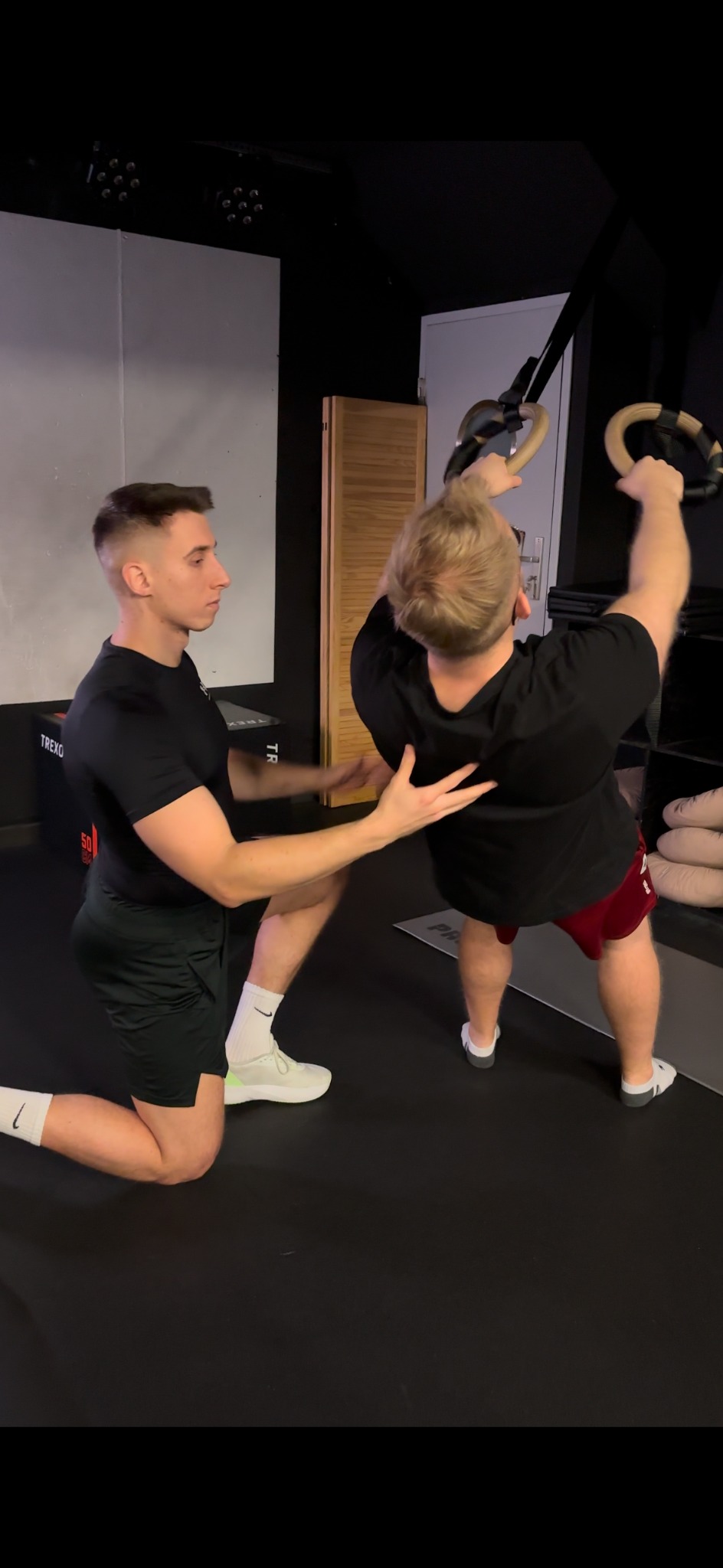 Trener asekuruje klienta podczas ćwiczeń na drążku gimnastycznym w siłowni. Trener w czarnej koszulce, klęcząc, pomaga ćwiczącemu. Ćwiczący tyłem do kamery.