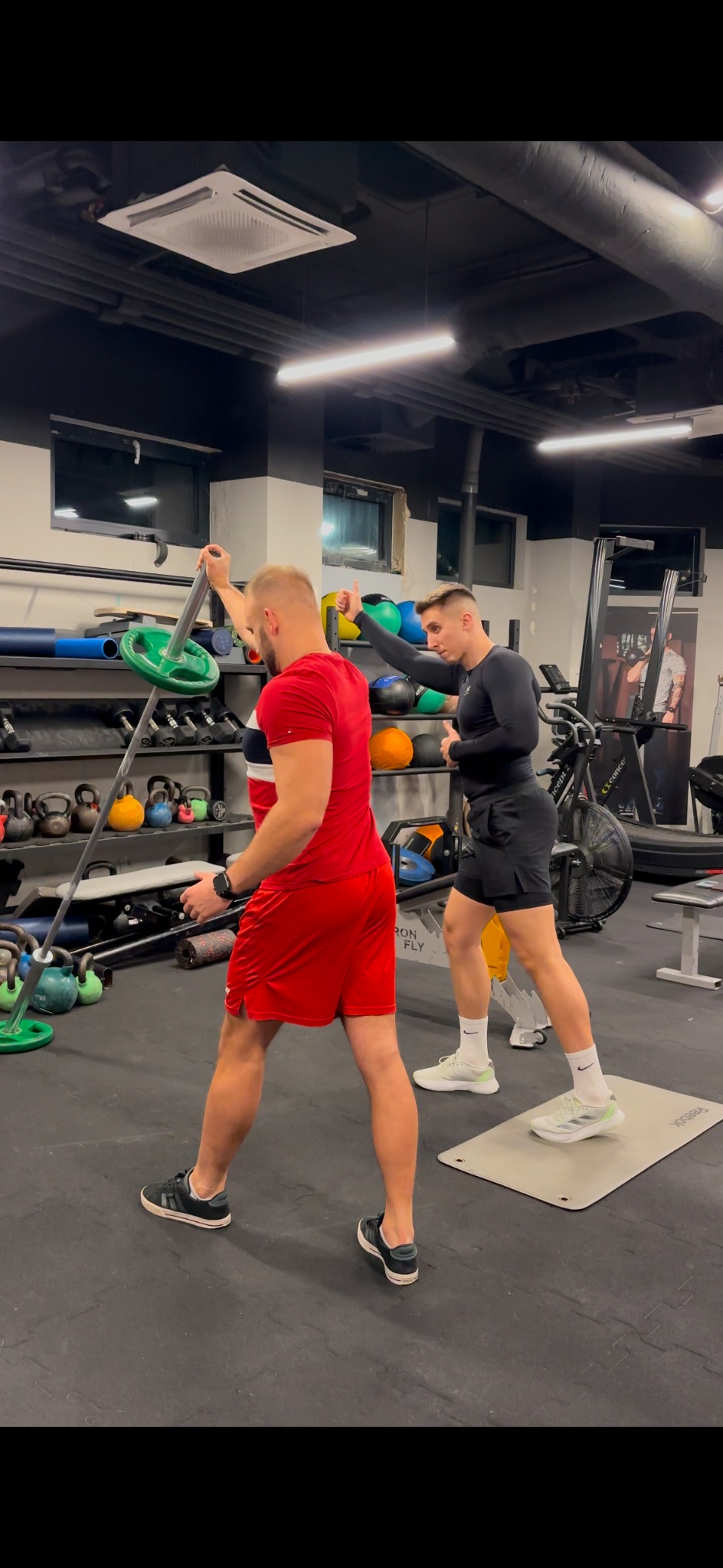 Trening z obciążeniem pod okiem instruktora w siłowni. Mężczyzna w czerwonej koszulce ćwiczy, a drugi instruuje, stojąc na macie. W tle sprzęt do ćwiczeń.