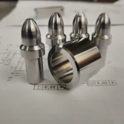 Precyzyjnie obrobione metalowe elementy, w tym cztery stożkowe trzpienie, z których dwa posiadają nacięcia, oraz pierścień, ułożone na tle technicznym z naniesionymi wymiarami i tolerancjami.