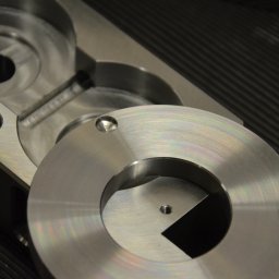 MECAMA SPÓŁKA Z OGRANICZONĄ ODPOWIEDZIALNOŚCIĄ - Dwa precyzyjnie obrobione, metalowe elementy z widocznymi otworami i gwintami, leżące na ciemnym, prążkowanym podłożu. Jeden element okrągły, drugi o bardziej złożonym kształcie.