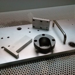 MECAMA SPÓŁKA Z OGRANICZONĄ ODPOWIEDZIALNOŚCIĄ - Metalowa płyta z uchwytami i różnymi elementami obrobionymi skrawaniem, w tym pierścień z otworem, prostopadłościan oraz element w kształcie litery T, na tle perforowanej powierzchni.