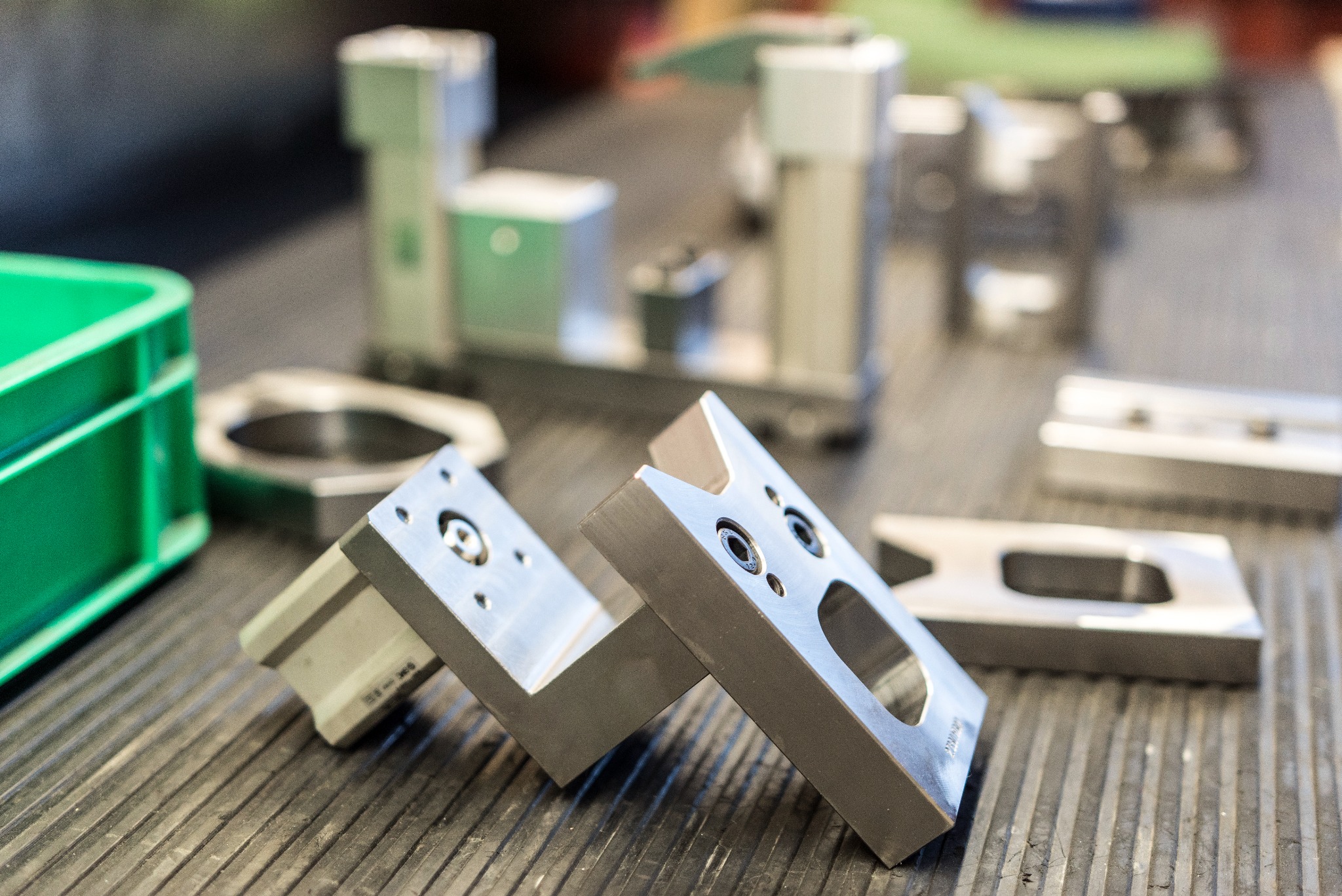 Precyzyjnie obrobione metalowe elementy z otworami i gwintami leżące na rowkowanym stole warsztatowym, w tle widoczne inne detale i zielony pojemnik.