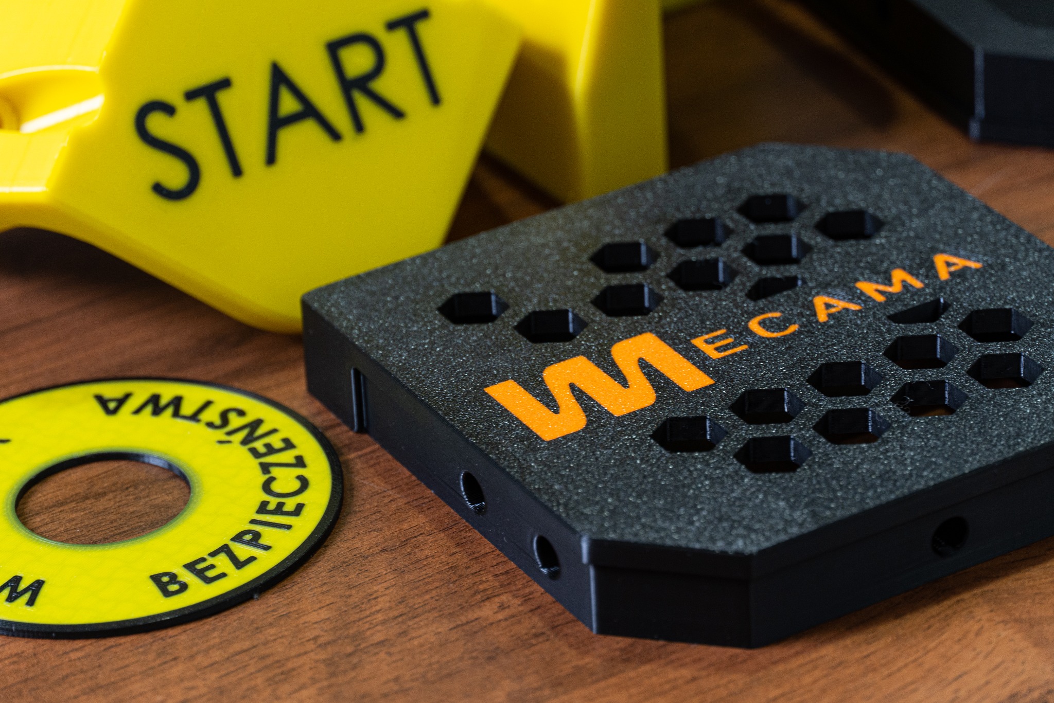 Czarny element z napisem 'MECAMA' wykonany w technologii druku 3D, leżący na drewnianym stole obok żółtych elementów z nadrukami 'START' i 'BEZPIECZEŃSTWA WŁASNE'.