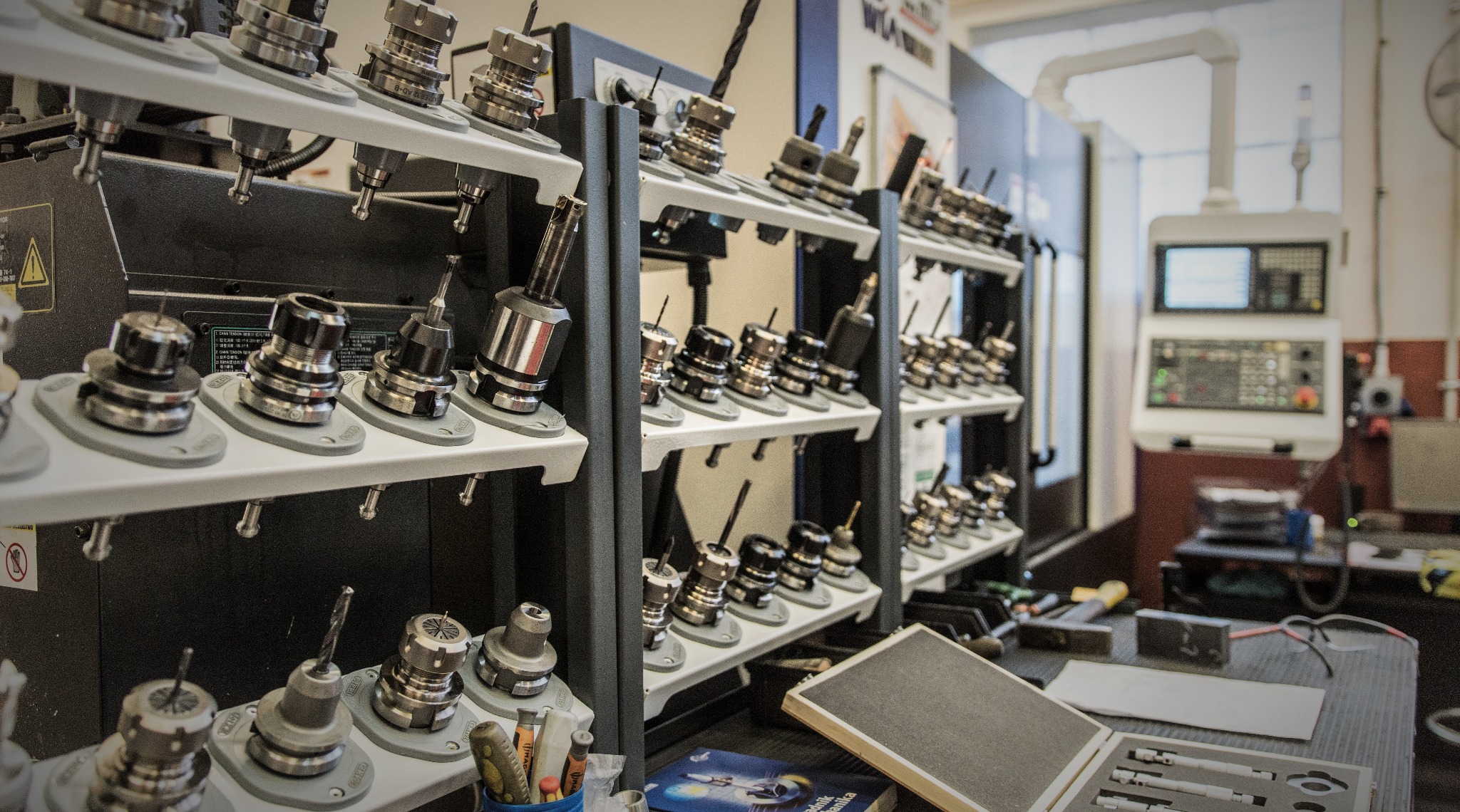 Rzędy uchwytów narzędziowych do obrabiarek CNC, uporządkowane na półkach w warsztacie, z widoczną maszyną CNC w tle i przyborami na stole roboczym.