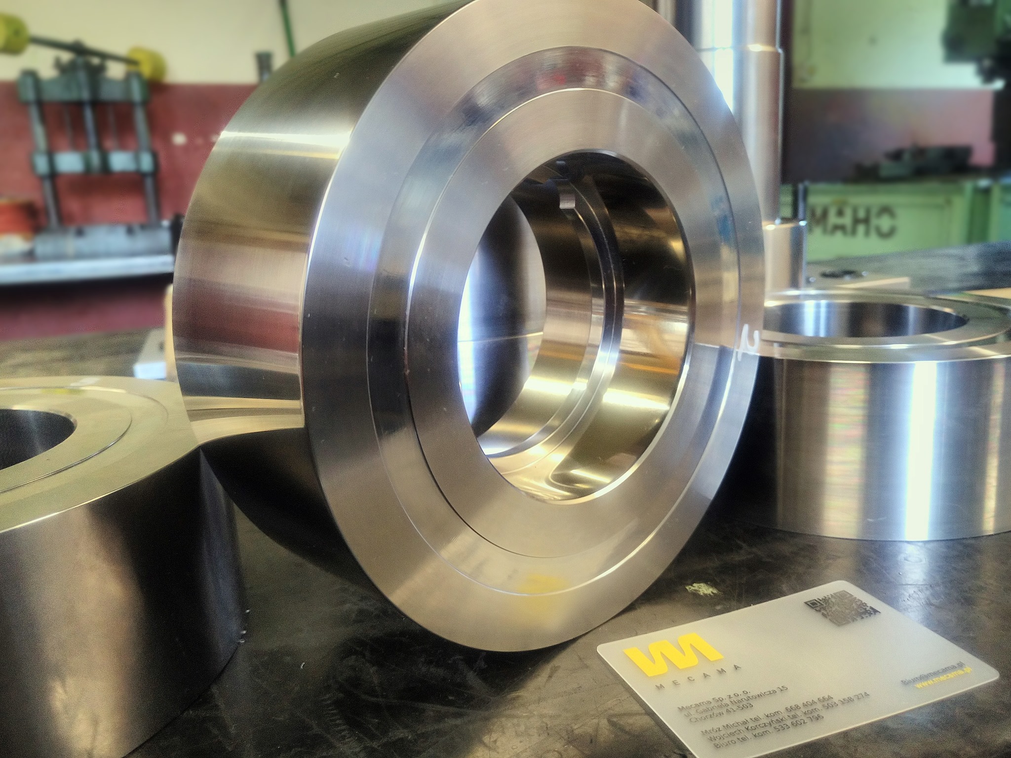 Precyzyjnie obrobiony, błyszczący metalowy pierścień z otworem, leżący na warsztatowym stole, obok wizytówka firmy Mecama i inne elementy obróbki, w tle maszyna MAHO.