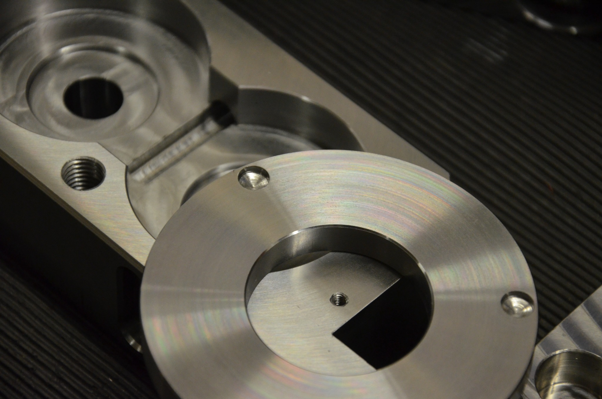 Dwa precyzyjnie obrobione, metalowe elementy z widocznymi otworami i gwintami, leżące na ciemnym, prążkowanym podłożu. Jeden element okrągły, drugi o bardziej złożonym kształcie.