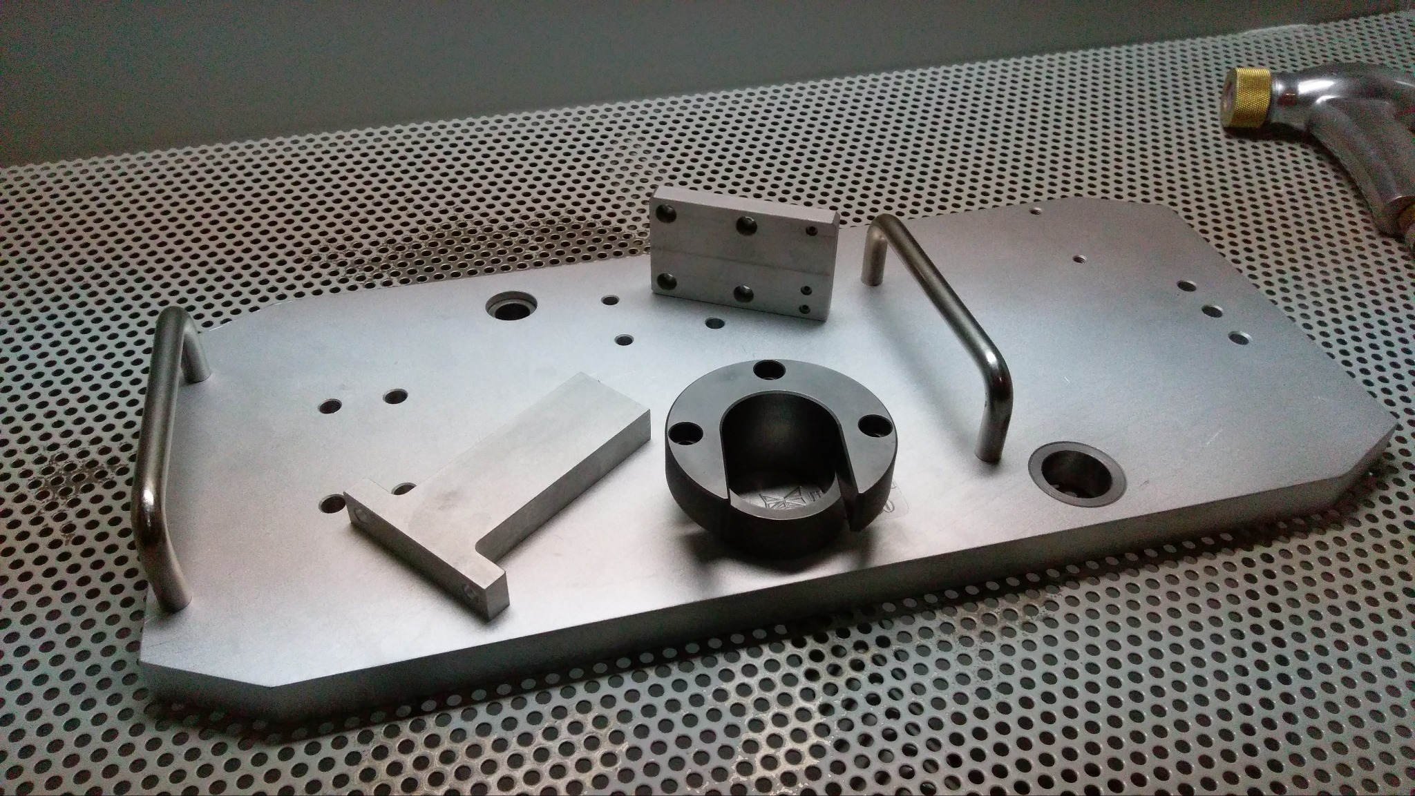 Metalowa płyta z uchwytami i różnymi elementami obrobionymi skrawaniem, w tym pierścień z otworem, prostopadłościan oraz element w kształcie litery T, na tle perforowanej powierzchni.