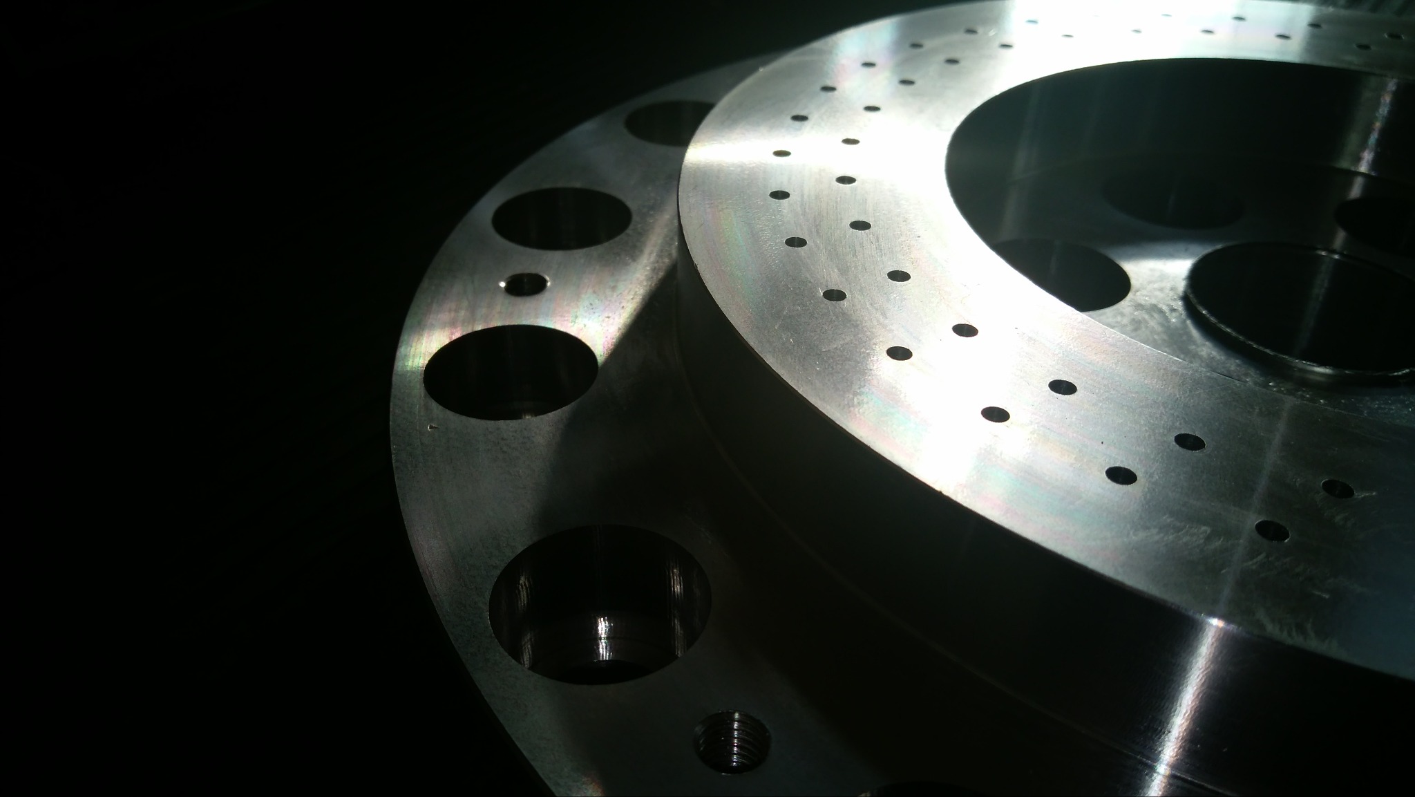 Dwa metalowe pierścienie z wywierconymi otworami, jeden z większymi otworami, drugi z mniejszymi, leżące jeden na drugim w ciemnym otoczeniu, oświetlone z boku, ukazujące metaliczny połysk...