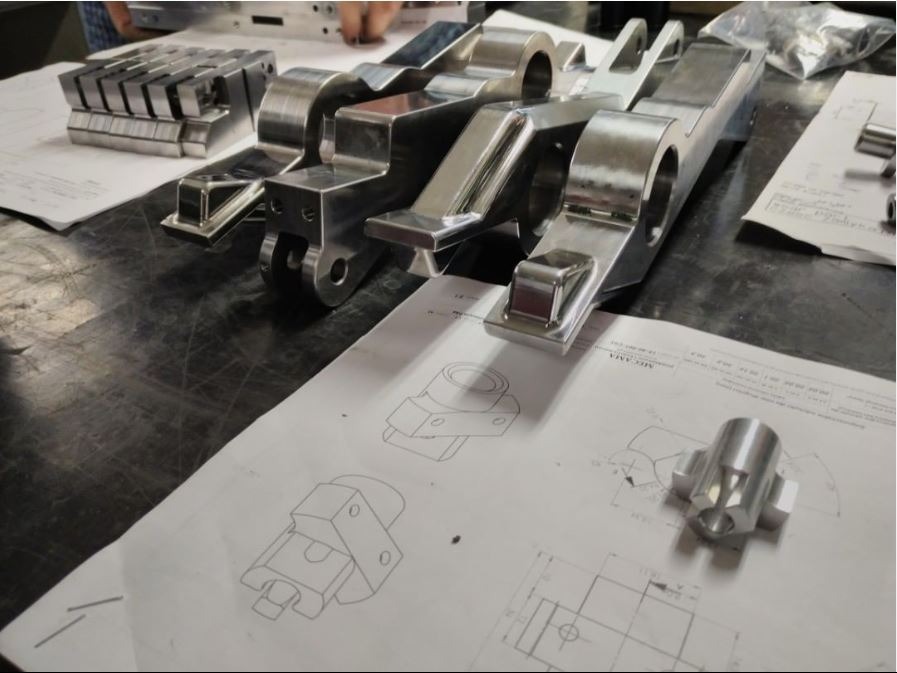 Błyszczące, metalowe elementy po obróbce, ułożone na stole obok rysunków technicznych z wymiarami.