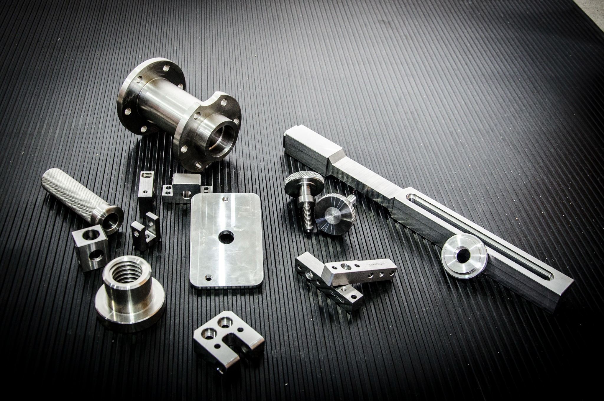 Zestaw precyzyjnie obrobionych, metalowych elementów o różnych kształtach i rozmiarach, ułożonych na czarnym, ryflowanym tle. Widoczne detale to m.in. kołnierz z otworami, gwintowane tuleje...