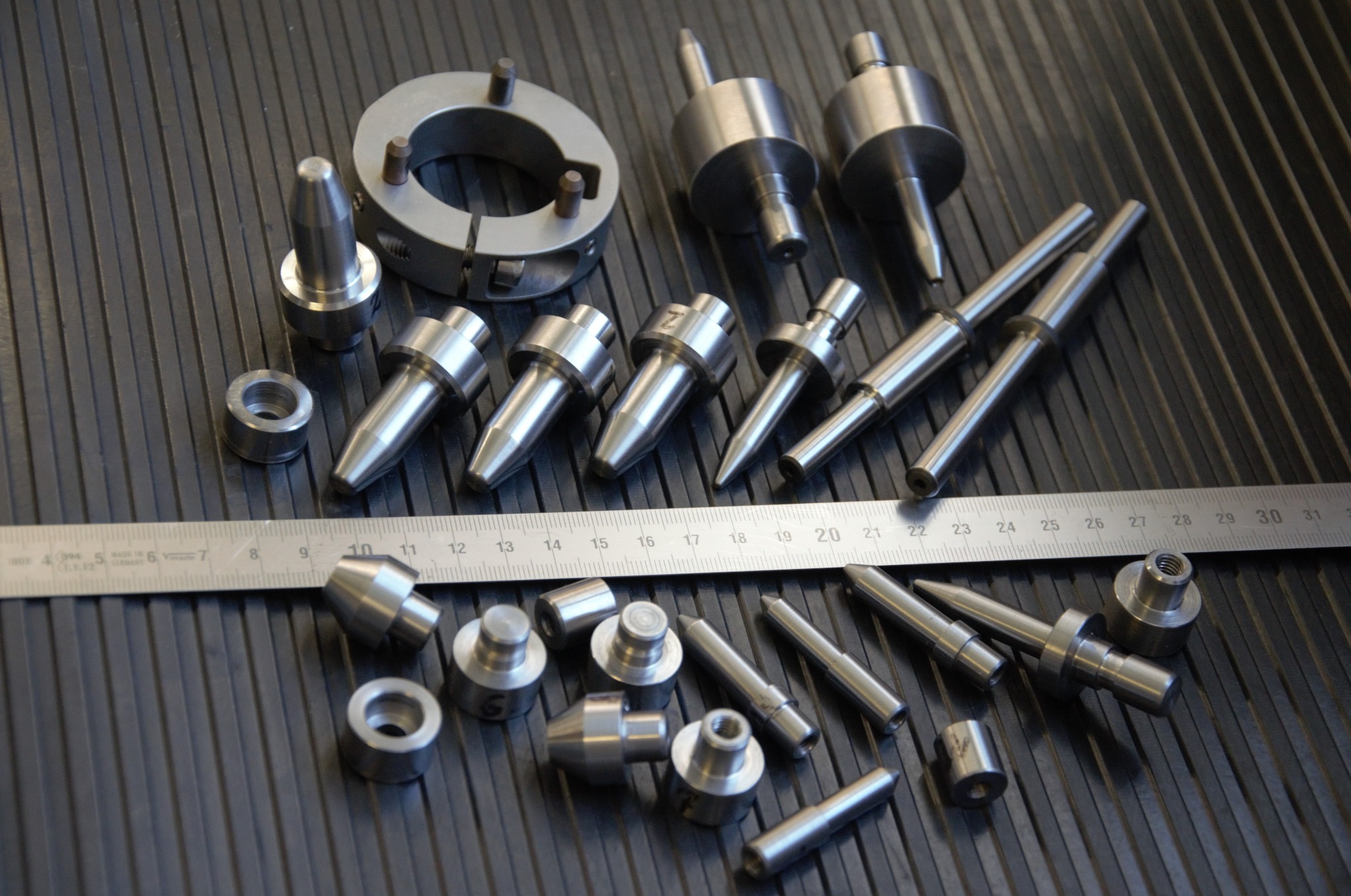 Zestaw precyzyjnie obrobionych, metalowych elementów o różnych kształtach, leżących na ciemnym, rowkowanym podłożu, z miarką w tle, ukazujący detale i dokładność wykonania.