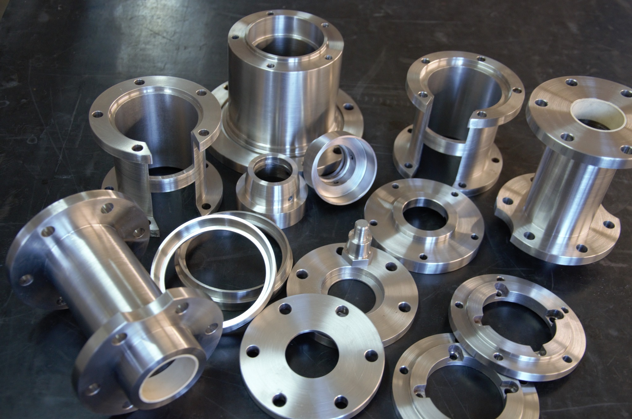 Zestaw precyzyjnie wykonanych, metalowych elementów o różnych kształtach i rozmiarach, w tym kołnierze, pierścienie i tuleje, z widocznymi śladami obróbki skrawaniem, ułożonych na ciemnej, matowej...