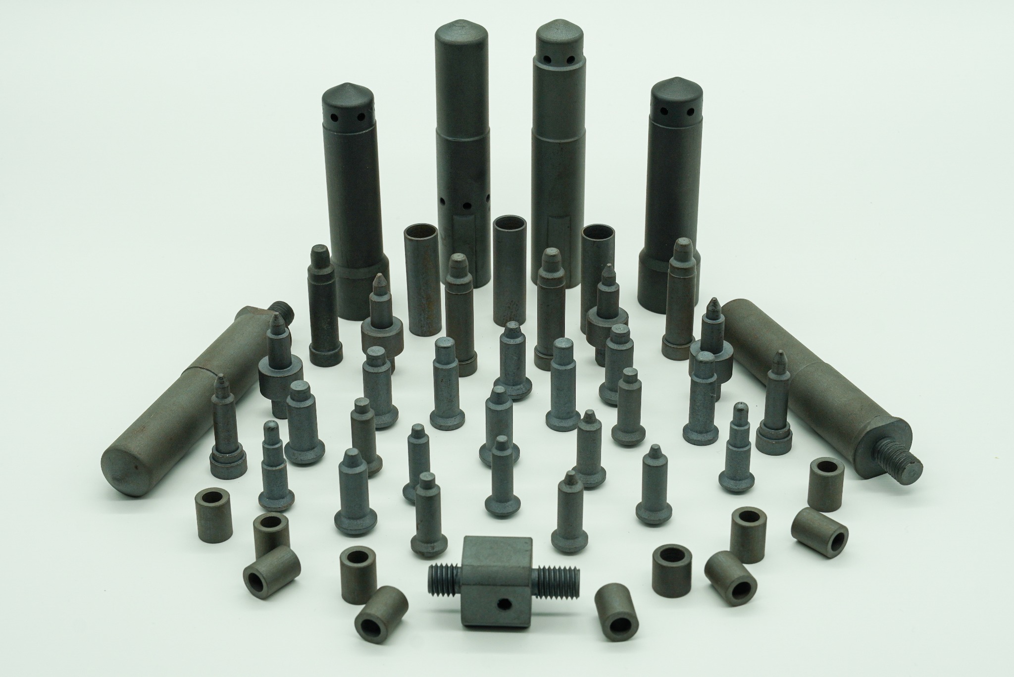 Zestaw precyzyjnie wykonanych, metalowych elementów o różnych kształtach i rozmiarach, ułożonych na białym tle, w tym cylindryczne trzpienie z otworami, tuleje i element z gwintem.