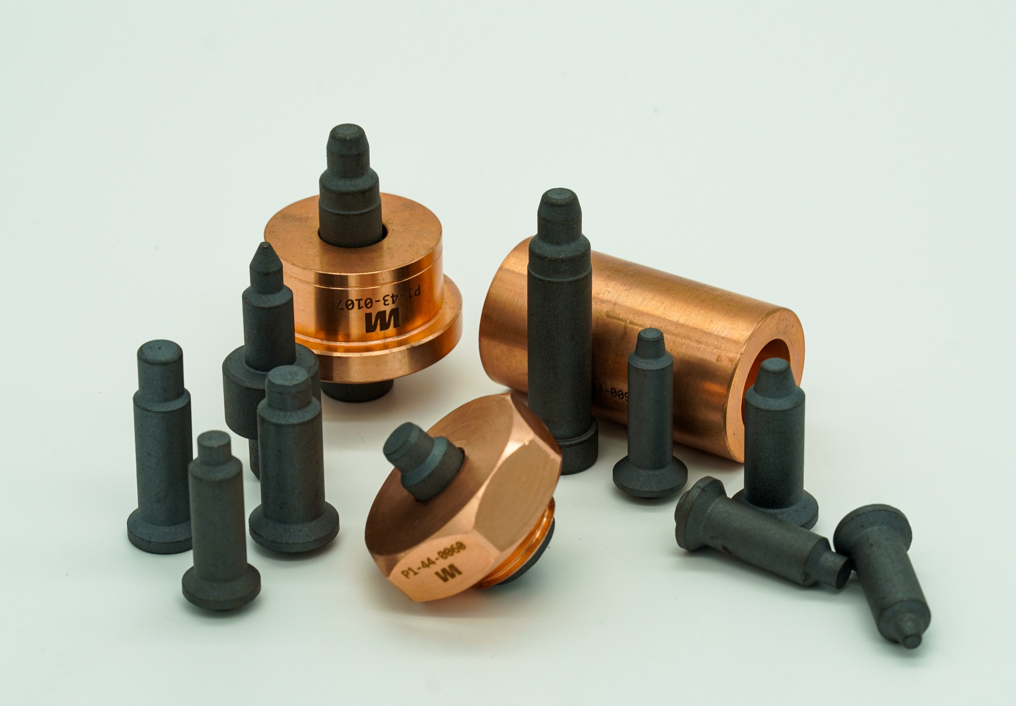 Precyzyjnie obrobione, metalowe elementy z miedzi i stali o różnych kształtach, w tym tuleje i trzpienie, ułożone na białym tle, widoczne oznaczenia kodowe na niektórych elementach.
