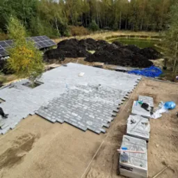 Fragment układanej szarej kostki na przygotowanym podłożu z widocznymi paletami z kostką, narzędziami i otoczeniem z drzewami, stawem oraz panelami słonecznymi.