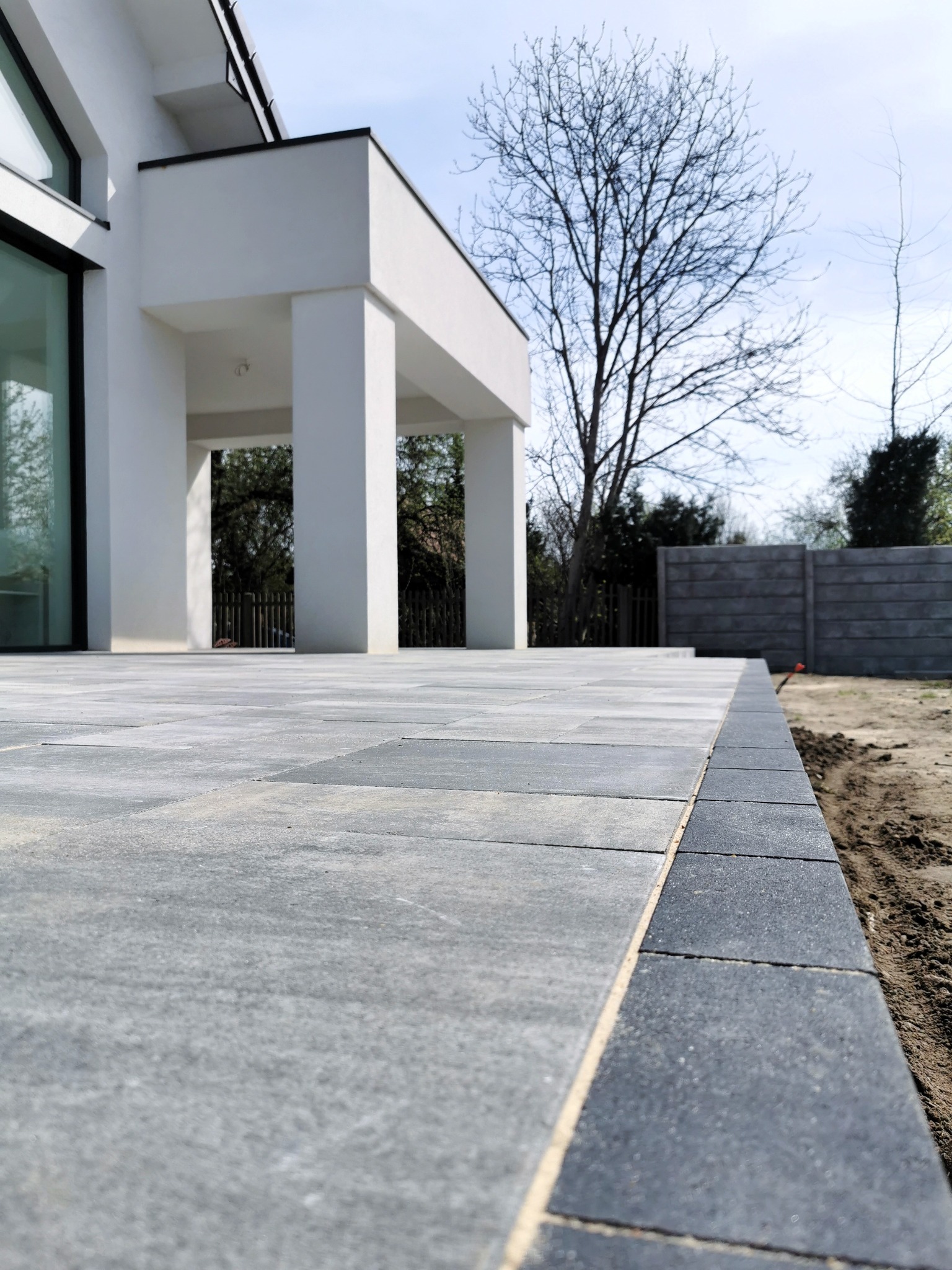 Nowoczesny taras z szarej kostki brukowej o minimalistycznym designie, z widocznym obrzeżem z ciemniejszej kostki. W tle biały budynek z dużymi oknami i drzewa bez liści na tle jasnego nieba.