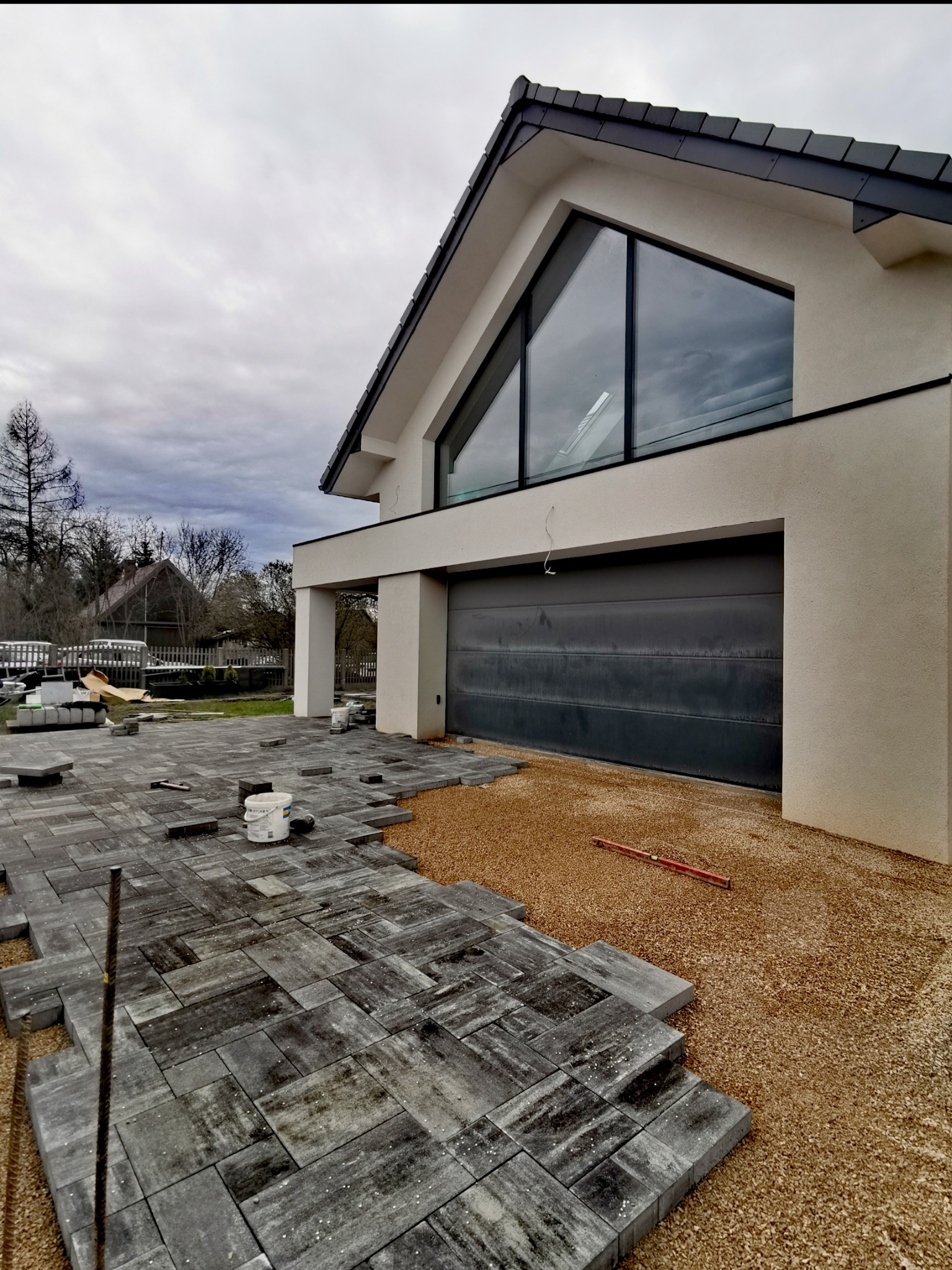 Częściowo ułożona, szara kostka na podsypce z piasku przed nowoczesnym domem z dużym oknem trójkątnym i garażem; widoczne narzędzia i wiadro.