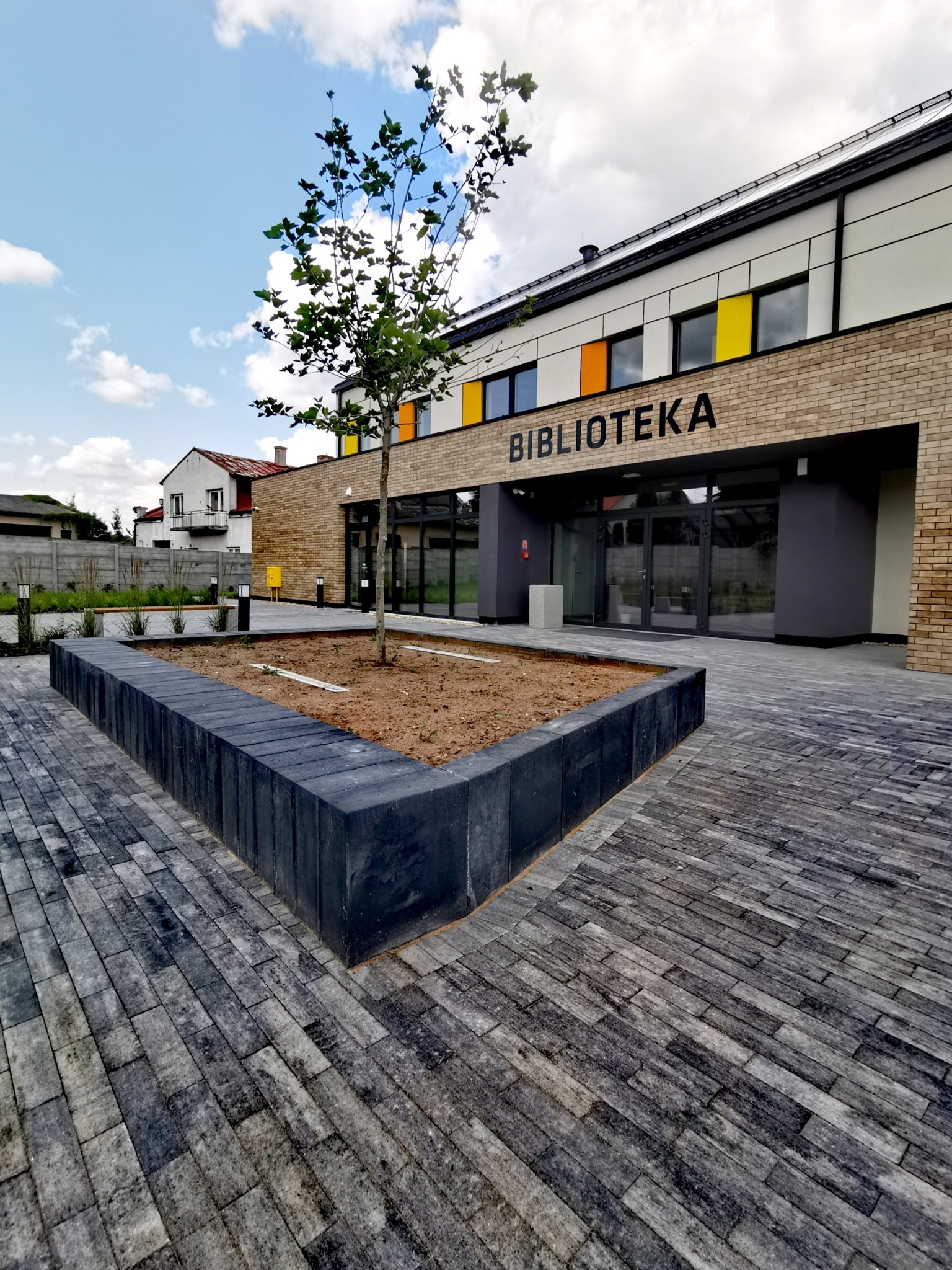 Nowoczesny budynek biblioteki z szarej cegły, z napisem 'BIBLIOTEKA' nad wejściem, otoczony geometrycznym klombem z młodym drzewkiem i ścieżką z szarej kostki brukowej ułożonej we wzór jodełki.