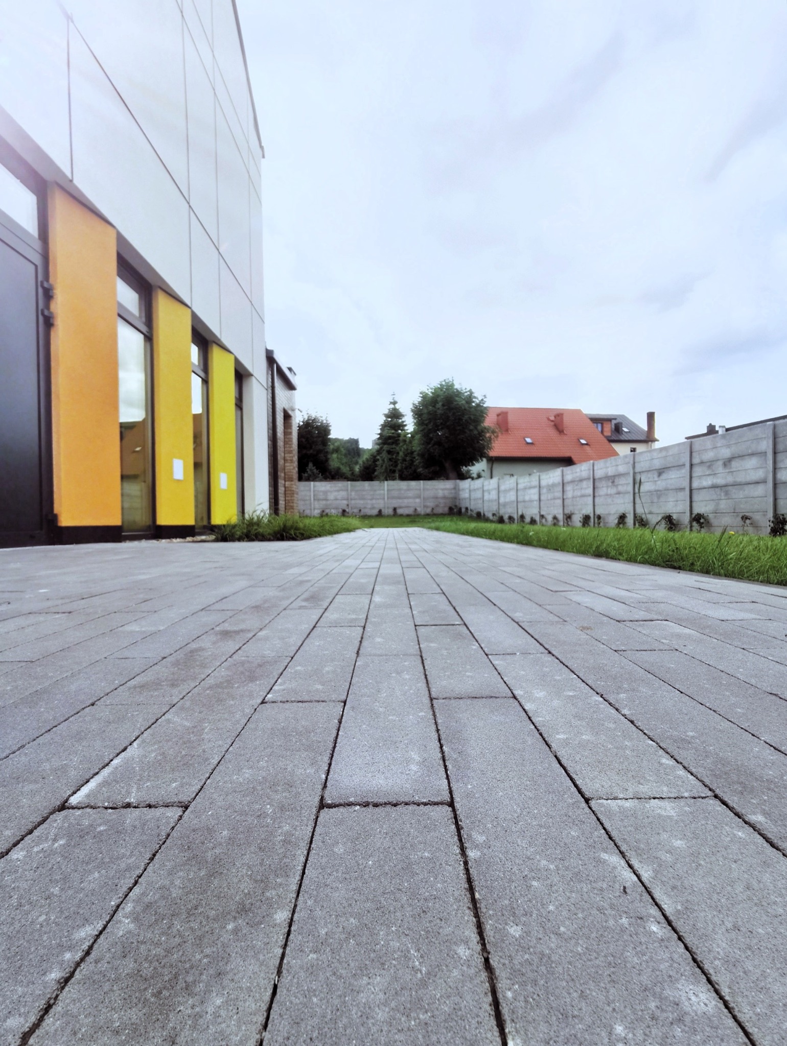 Ułożona szara kostka brukowa prowadząca wzdłuż nowoczesnego budynku z kolorowymi akcentami, w tle betonowe ogrodzenie i dom z czerwonym dachem.