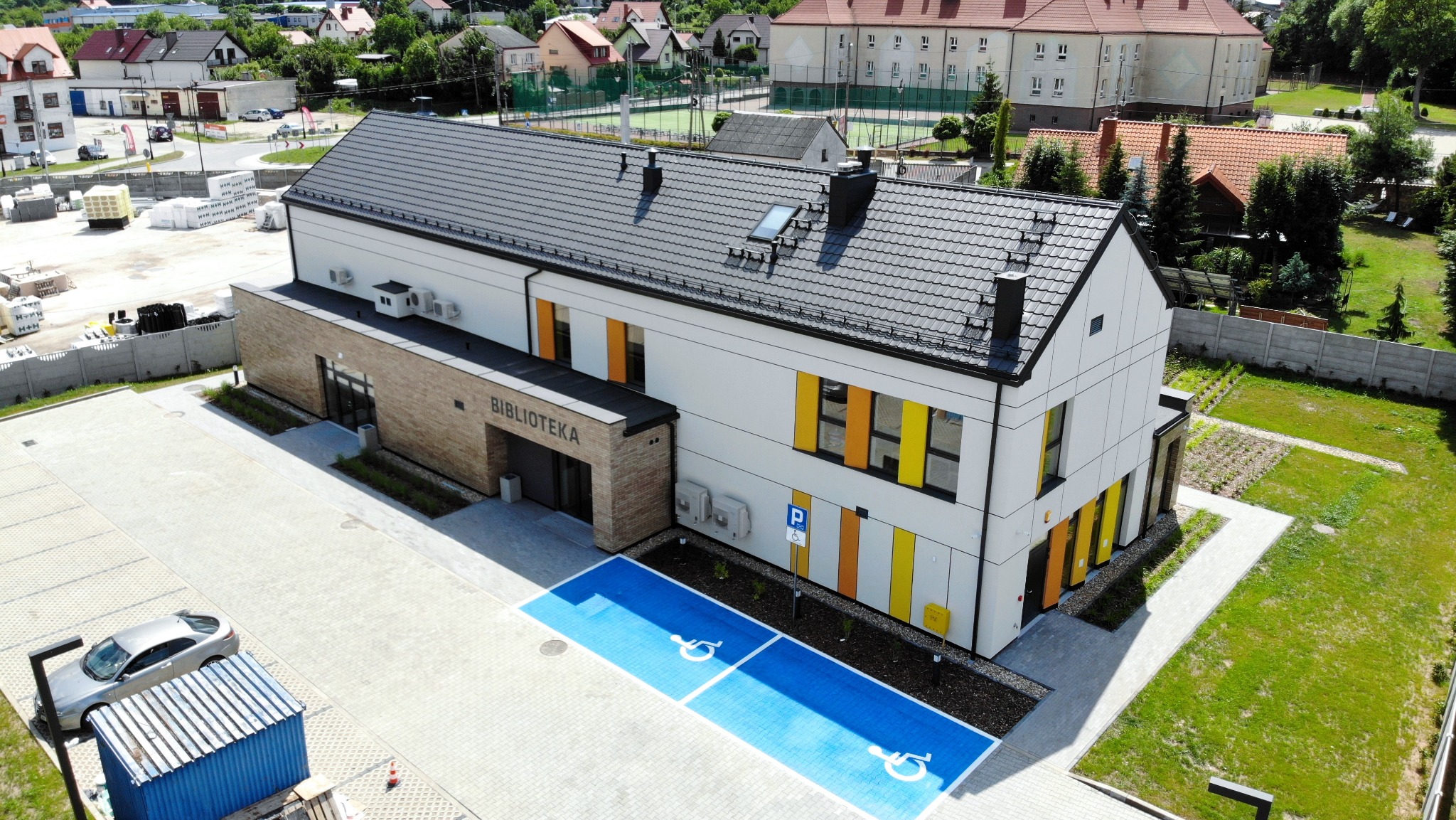 Nowoczesny budynek biblioteki z jasną elewacją, grafitowym dachem i kolorowymi akcentami okiennymi, widok z góry na parking z wyznaczonymi miejscami dla osób niepełnosprawnych, otoczony zielenią...