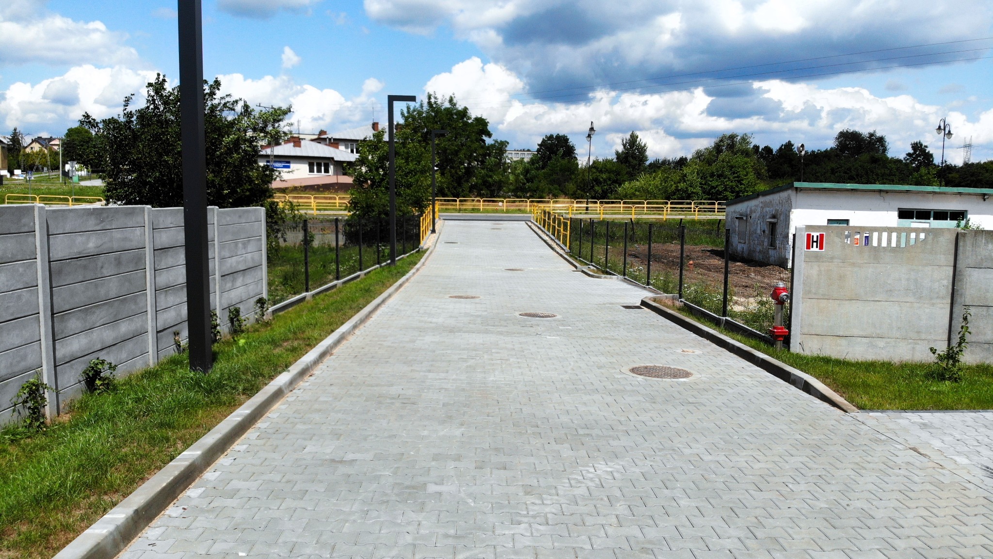 Szeroka, szara nawierzchnia z kostki brukowej w jodełkę, otoczona betonowymi krawężnikami i żółtymi barierkami, prowadząca do budynku na tle błękitnego nieba z chmurami.