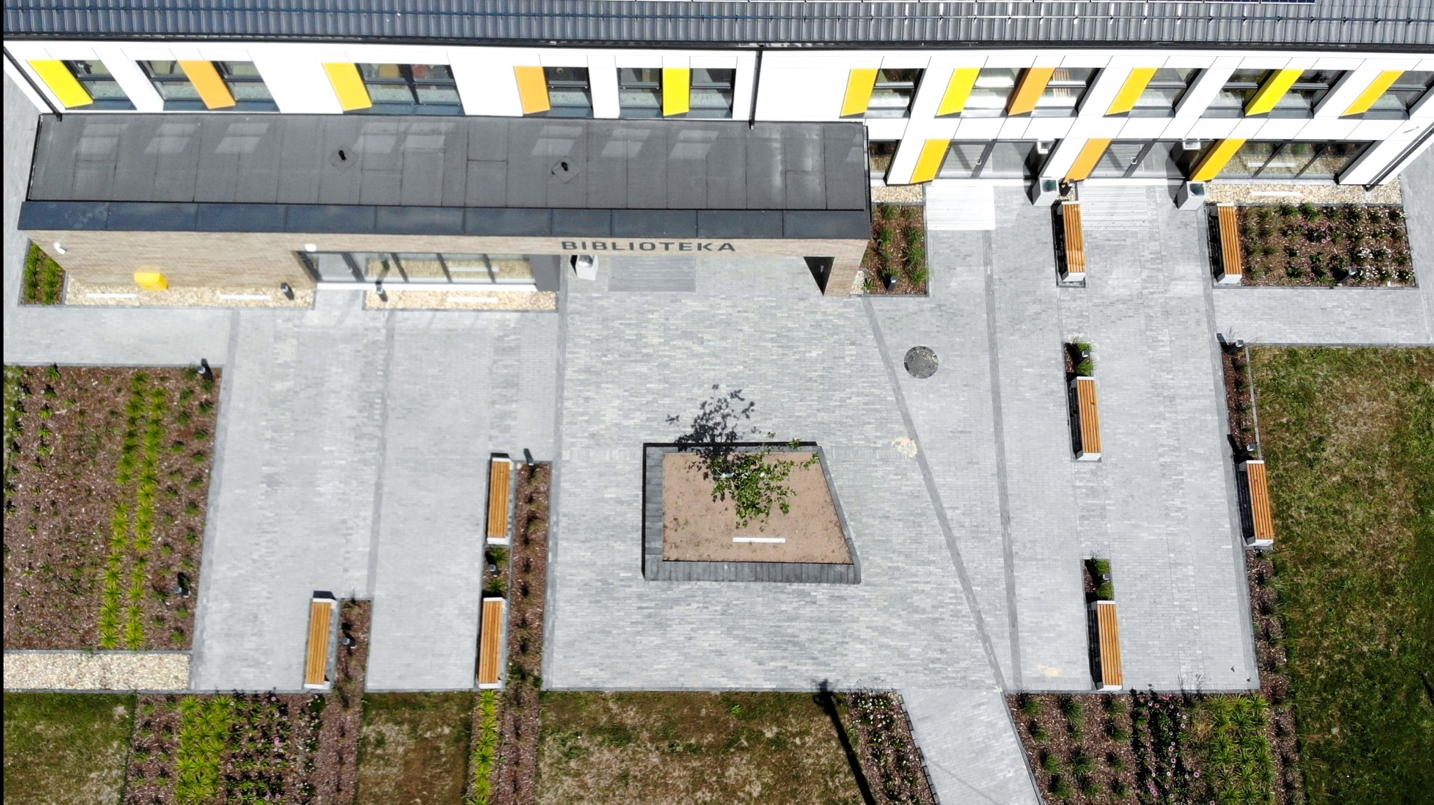 Widok z góry na nowo wybrukowany plac przed budynkiem Biblioteki z szarej kostki brukowej, z symetrycznie rozmieszczonymi ławkami, rabatami kwiatowymi i trawnikami. Centralnie umieszczona rabata...