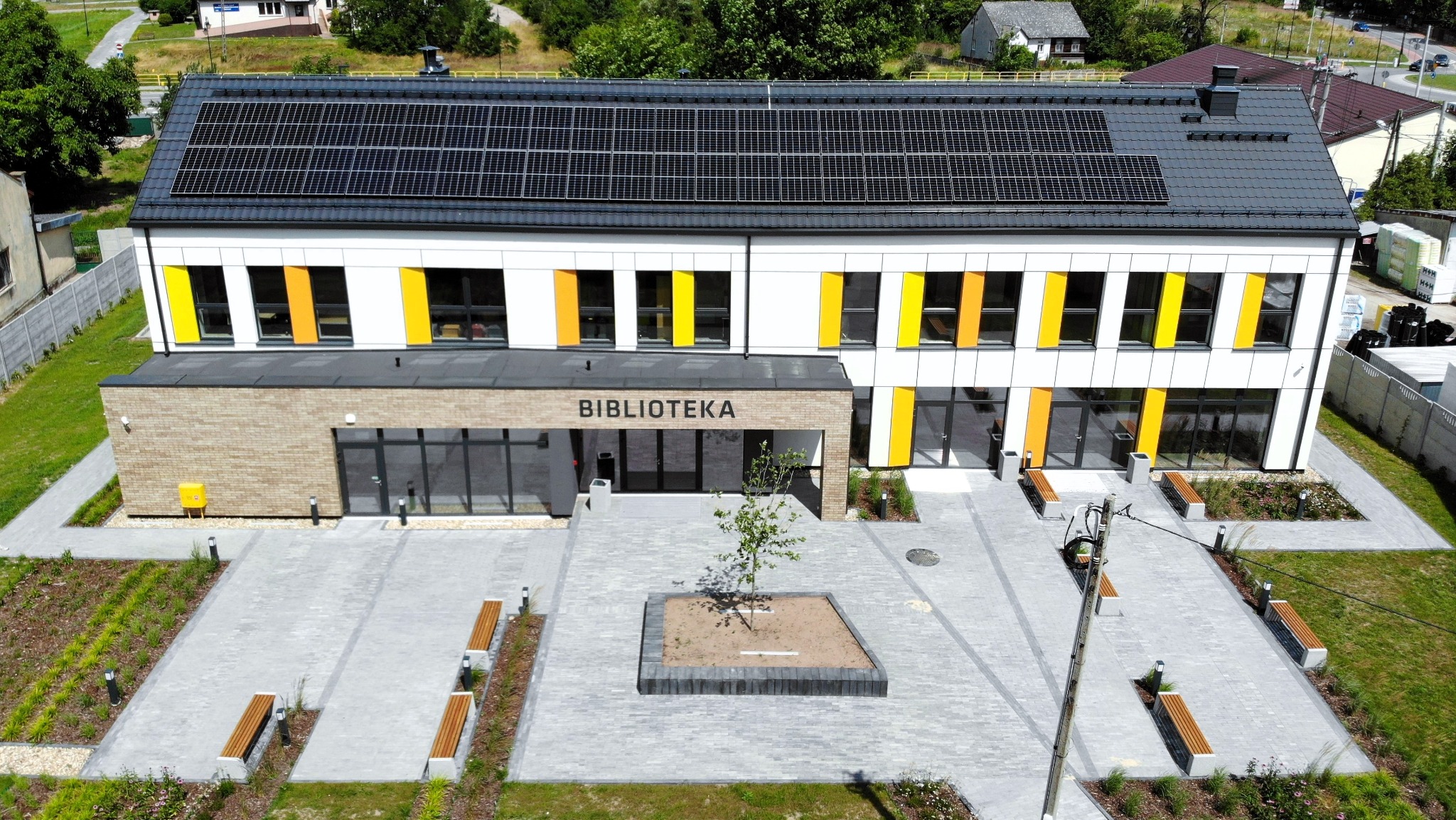 Nowoczesny budynek biblioteki z panelami słonecznymi na dachu, otoczony szarą kostką brukową, ławkami i młodym drzewkiem w centralnym punkcie placu przed wejściem.