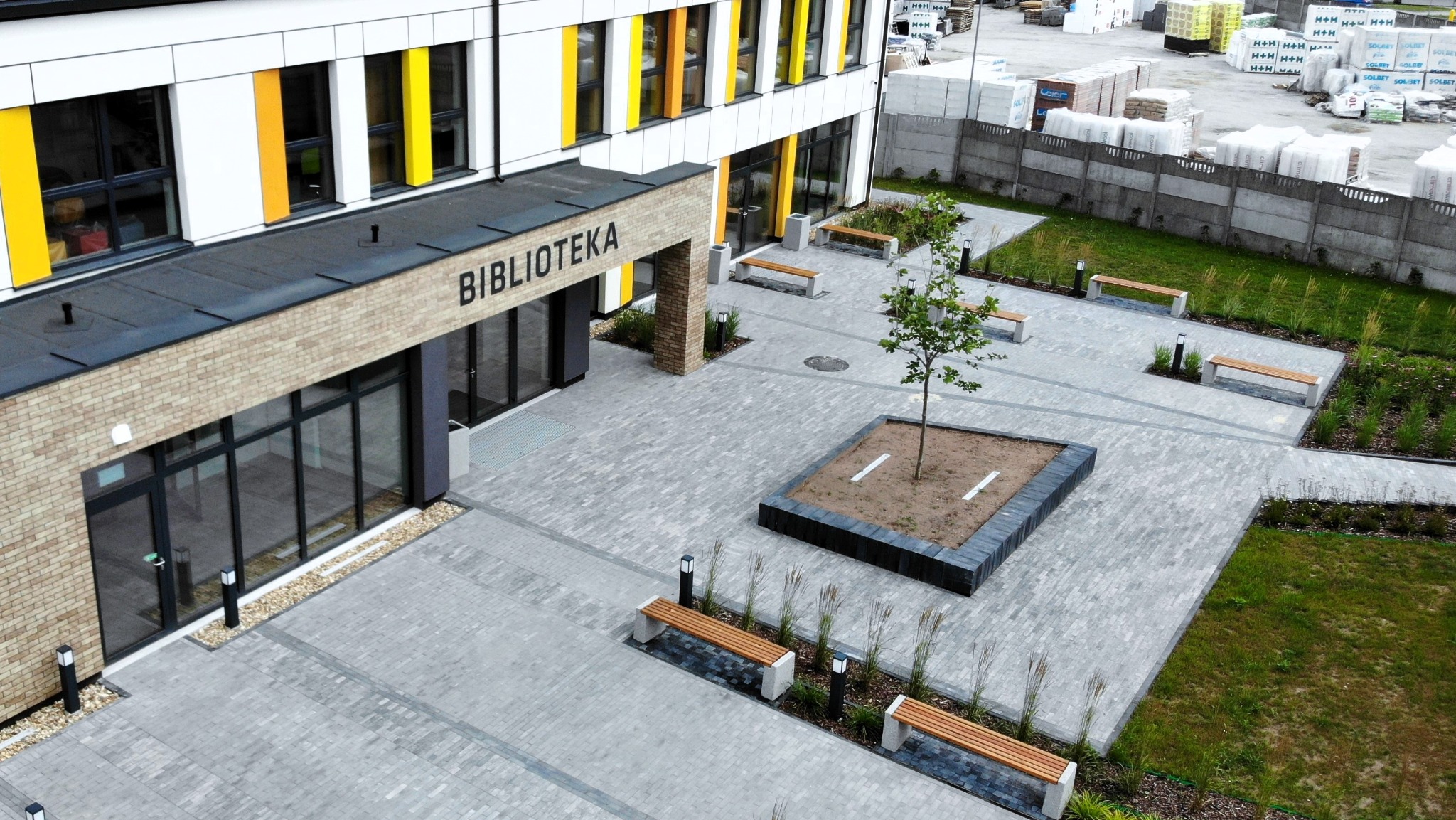 Nowoczesny plac przed budynkiem biblioteki, wyłożony szarą kostką brukową w różnych odcieniach, z elementami małej architektury: ławkami z drewnianymi siedziskami, niskimi lampami i rabatami...