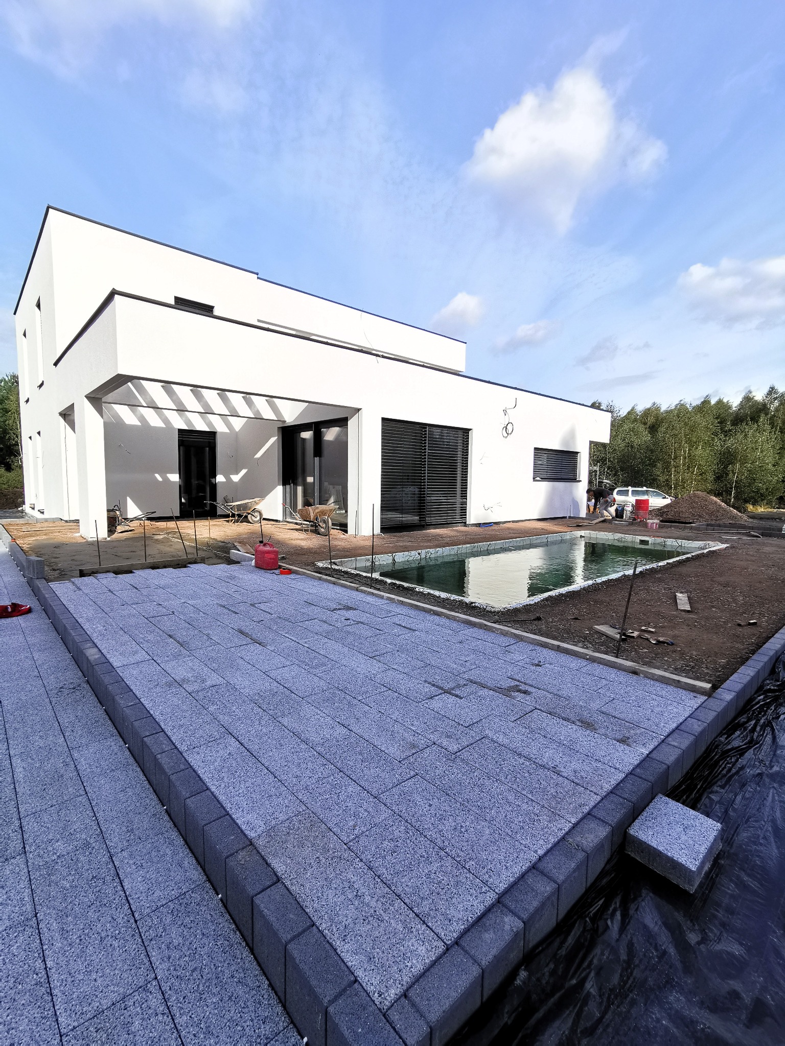 Nowoczesny dom jednorodzinny z szarej kostki brukowej, częściowo ułożonej, obok basenu w trakcie budowy, w tle drzewa i niebieskie niebo.