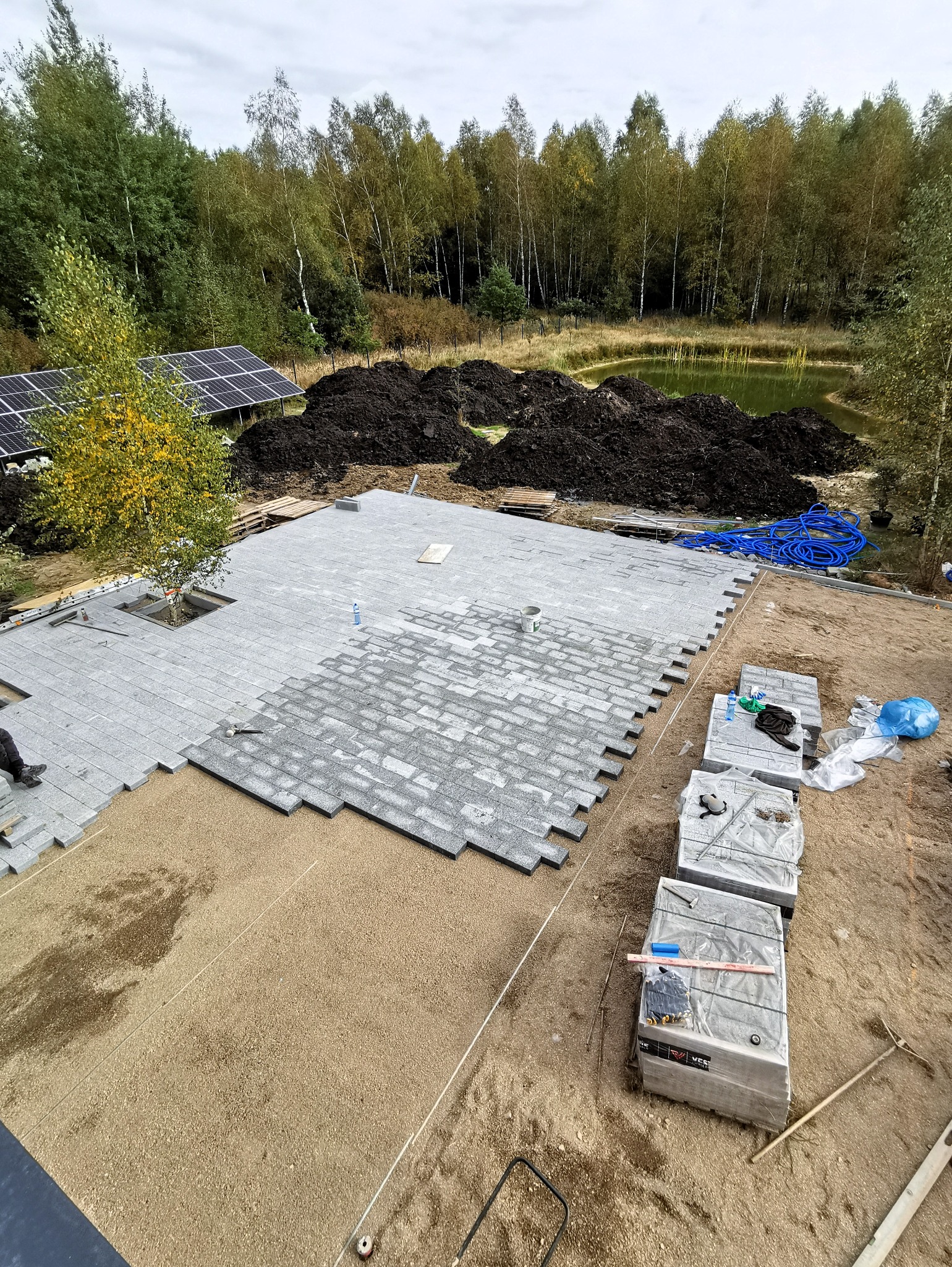 Fragment układanej szarej kostki na przygotowanym podłożu z widocznymi paletami z kostką, narzędziami i otoczeniem z drzewami, stawem oraz panelami słonecznymi.