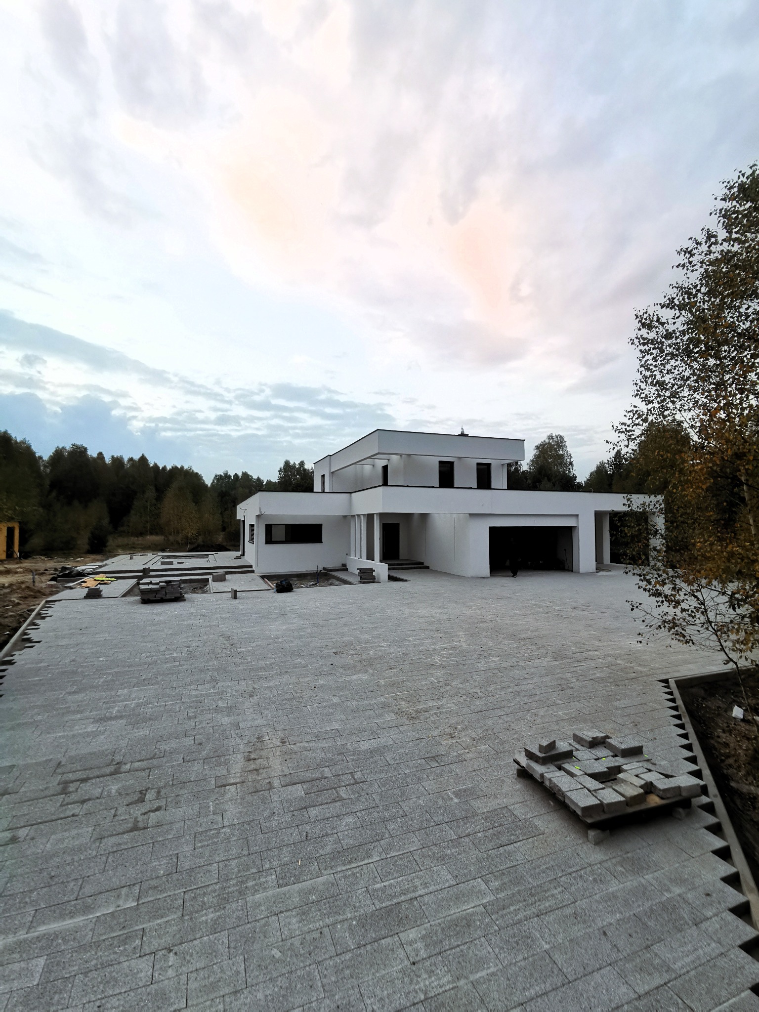 Nowoczesny dom jednorodzinny z płaskim dachem i jasną elewacją, otoczony rozległym podjazdem z szarej kostki, z paletą kostki przygotowanej do dalszych prac, widoczne drzewa w tle pod pochmurnym...