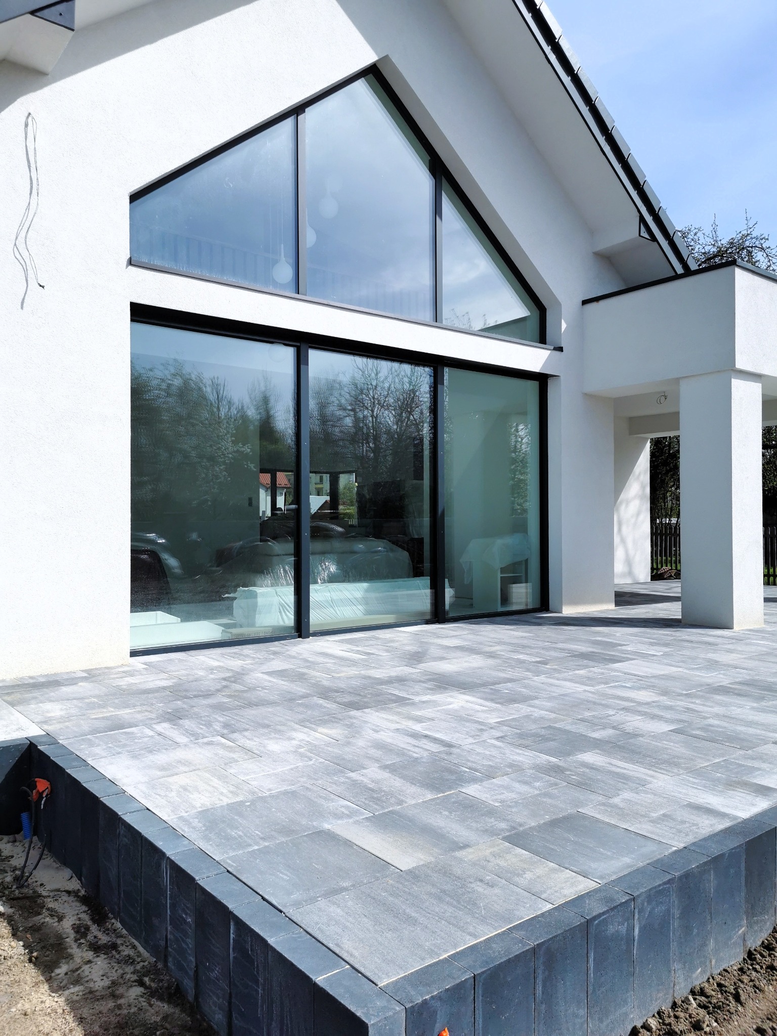 Nowoczesny taras w Kielcach wyłożony szarą kostką brukową, obrzeża z ciemnej kostki. Widok na biały budynek z dużymi oknami.