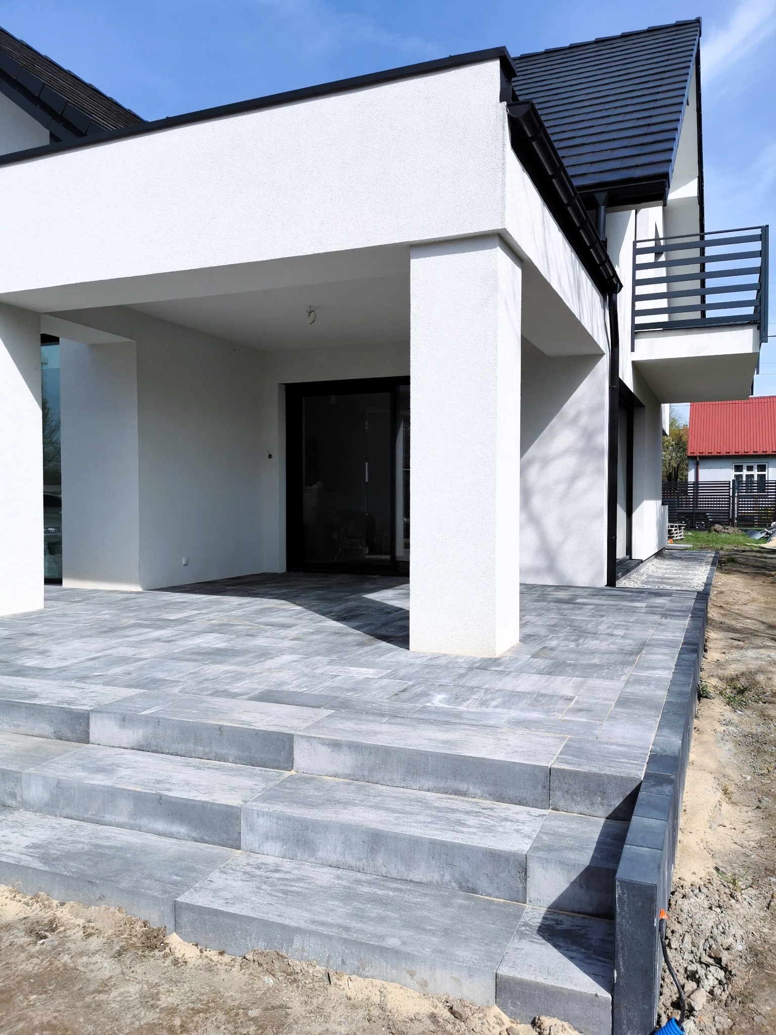 Nowoczesny dom z tarasem wyłożonym szarą kostką, widoczne schody z kostki prowadzące do wejścia, jasna elewacja budynku i czarny dach.