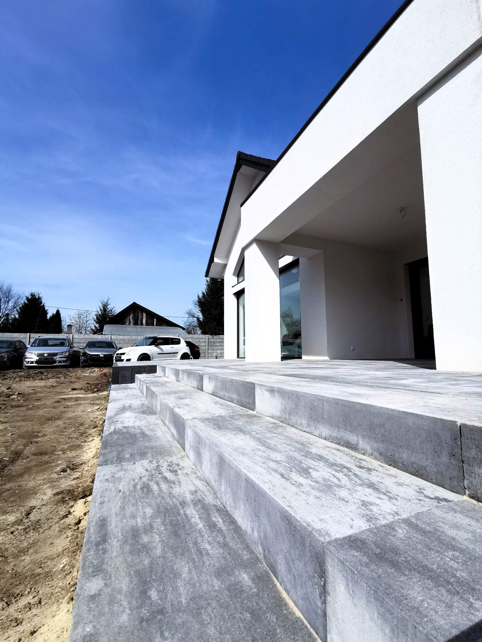 Nowoczesne schody wejściowe z szarej kostki brukowej prowadzące do białego domu z płaskim dachem, widok z dołu, w tle zaparkowane samochody i błękitne niebo.