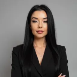 Portret kobiety w czarnej marynarce z delikatnym naszyjnikiem na szarym tle, skrzyżowane ręce.