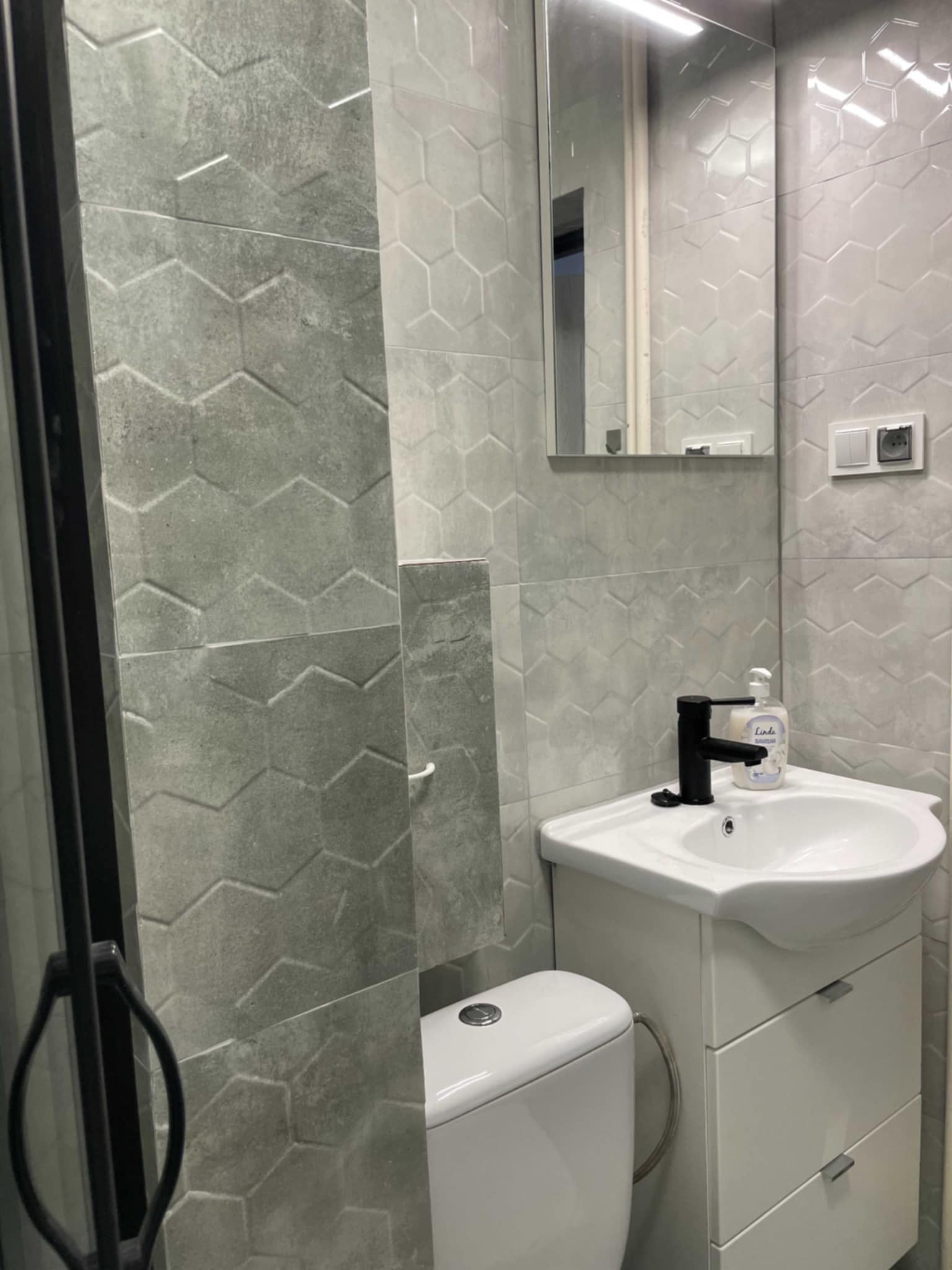 Szara łazienka z geometrycznymi płytkami, czarną armaturą i białą szafką pod umywalką, widok na toaletę i fragment kabiny prysznicowej.