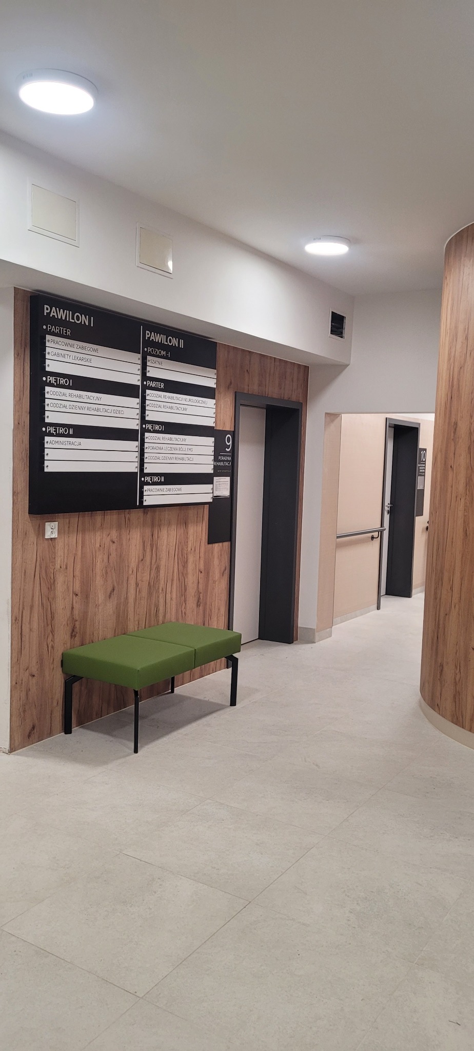 Korytarz w budynku medycznym z drewnianymi panelami ściennymi, tablicą informacyjną z nazwami gabinetów, zieloną ławką i szarą podłogą z płytek.